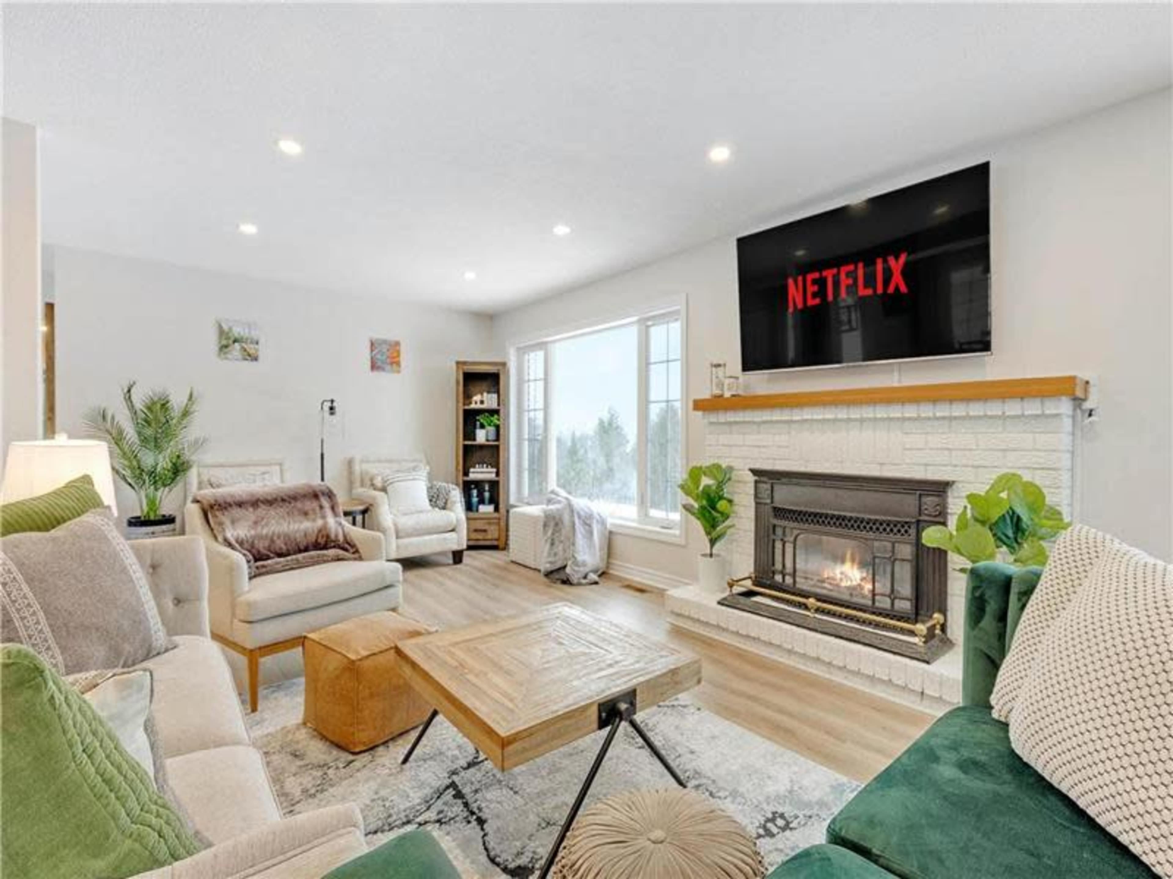 A modern living room features a fireplace, a large television displaying Netflix, and comfortable seating arrangements with natural light streaming through the windows.