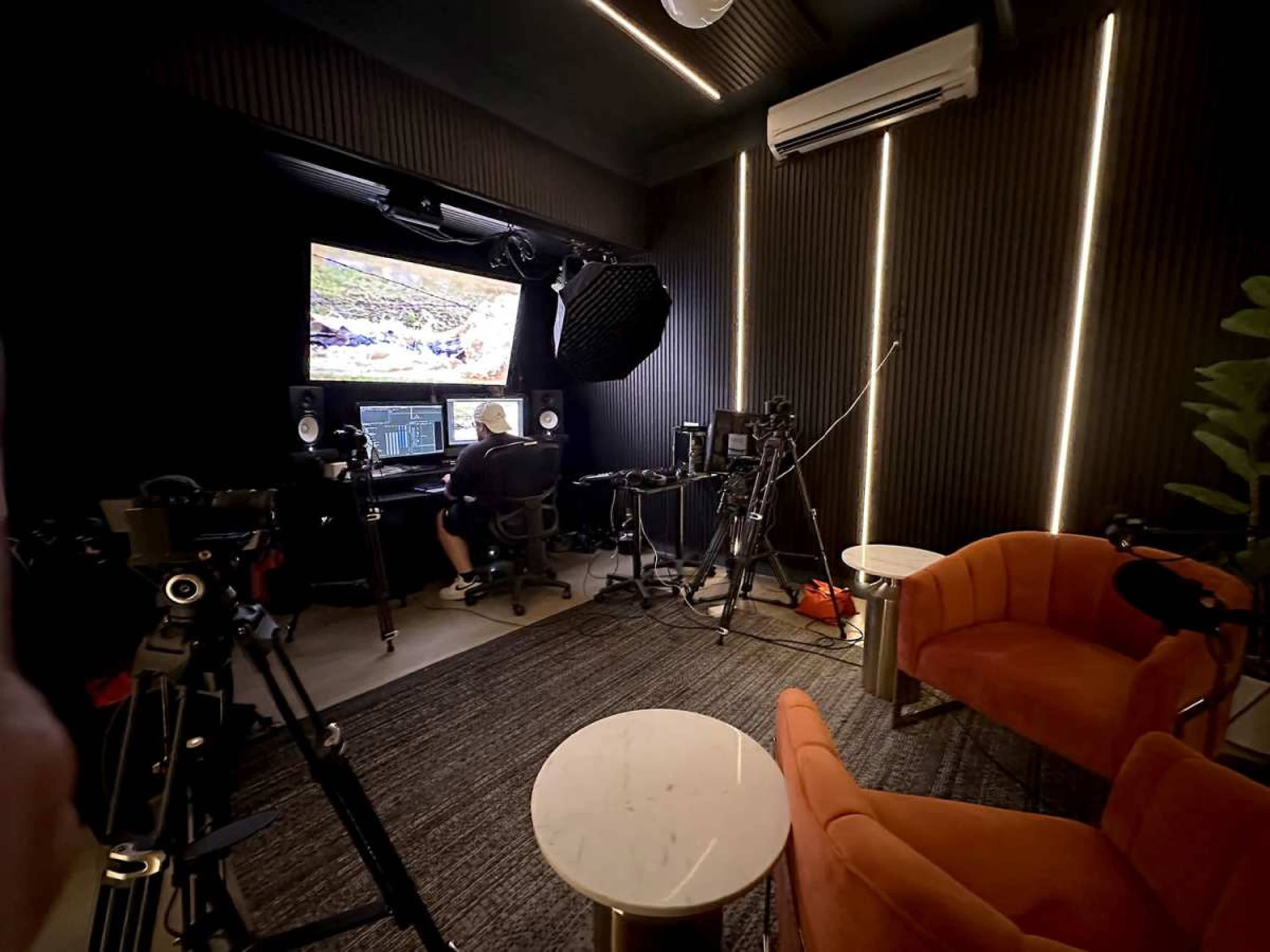 A person is seated at a desk with monitors in a modern studio equipped with cameras and orange chairs.