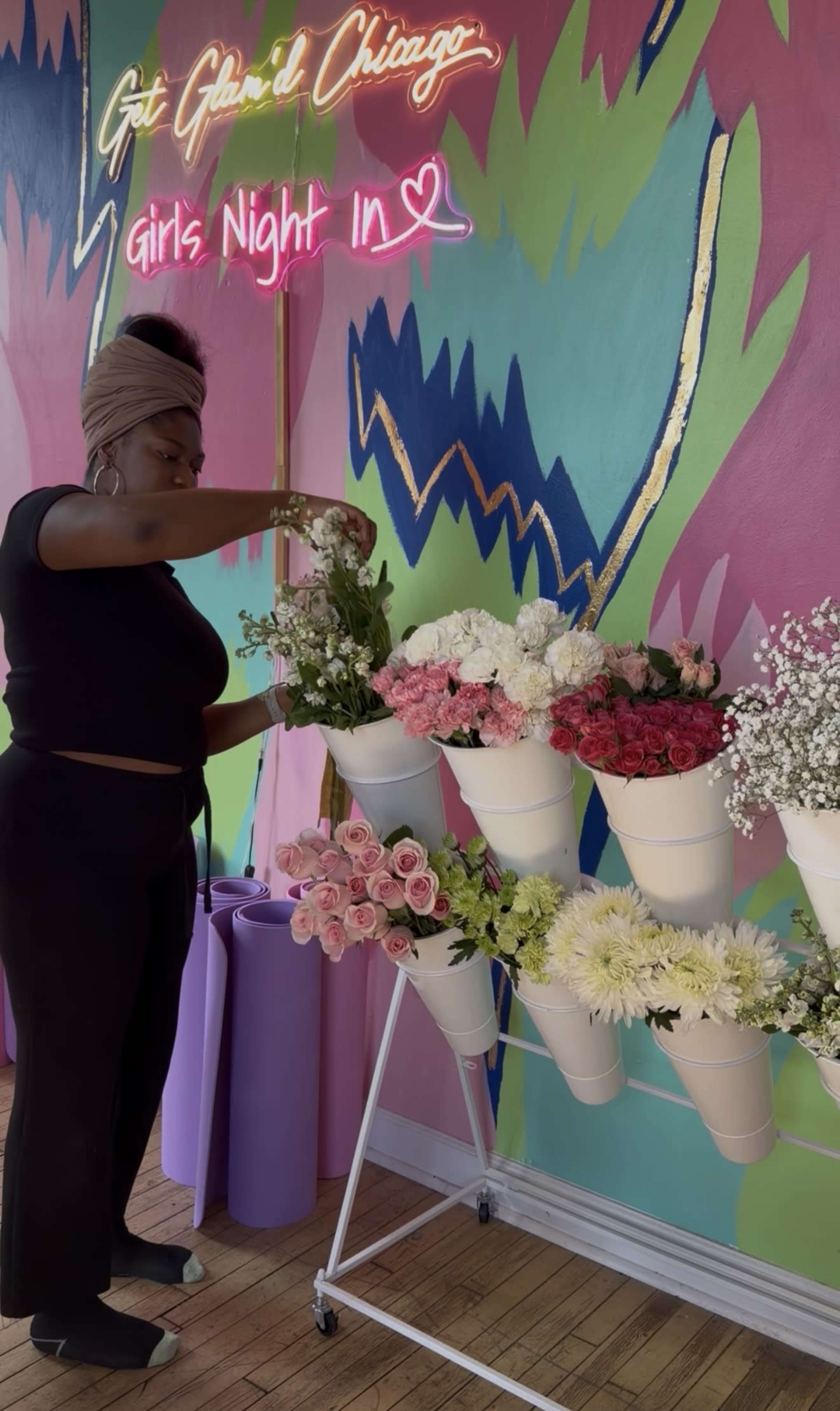 A woman arranges flowers in white pots on a display stand against a colorful mural.