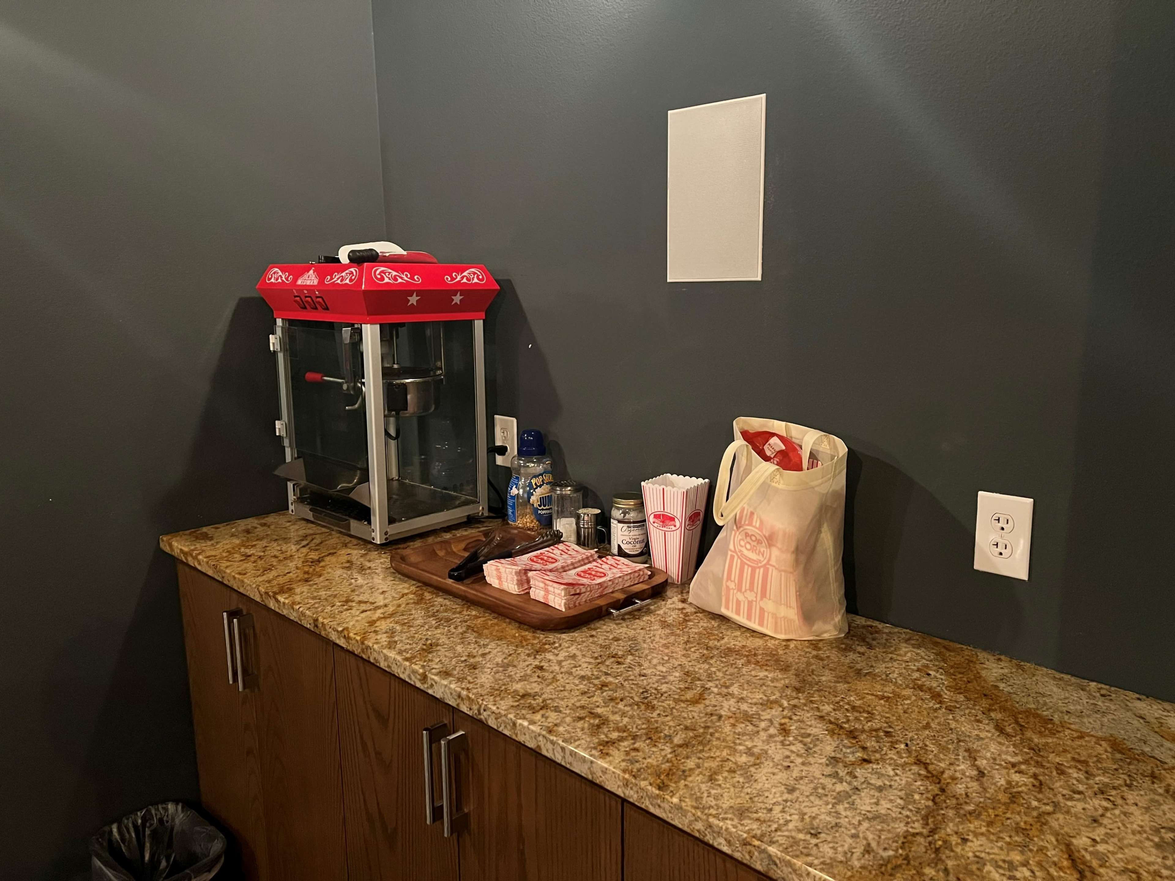 A countertop features a popcorn machine, condiments, disposable cups, and a tray of snacks on a wooden surface against a dark wall.