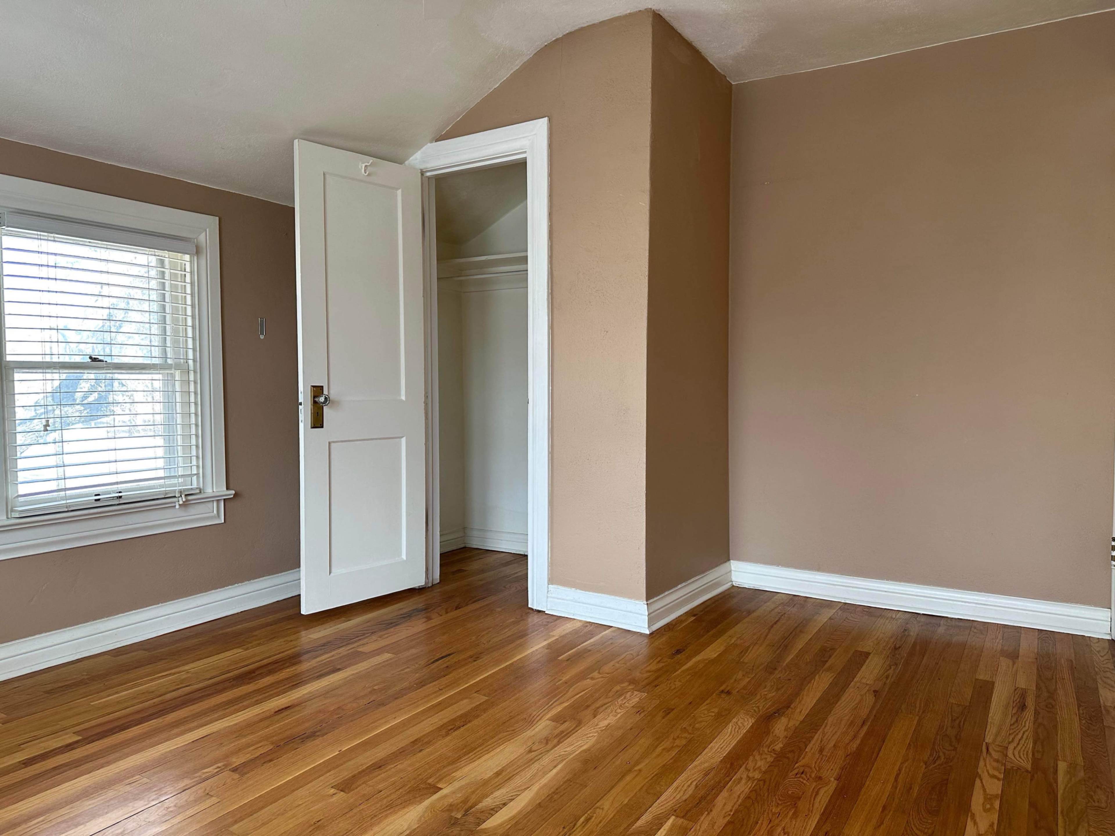A room with hardwood floors, beige walls, a window with white blinds, and an open door leading to a closet.