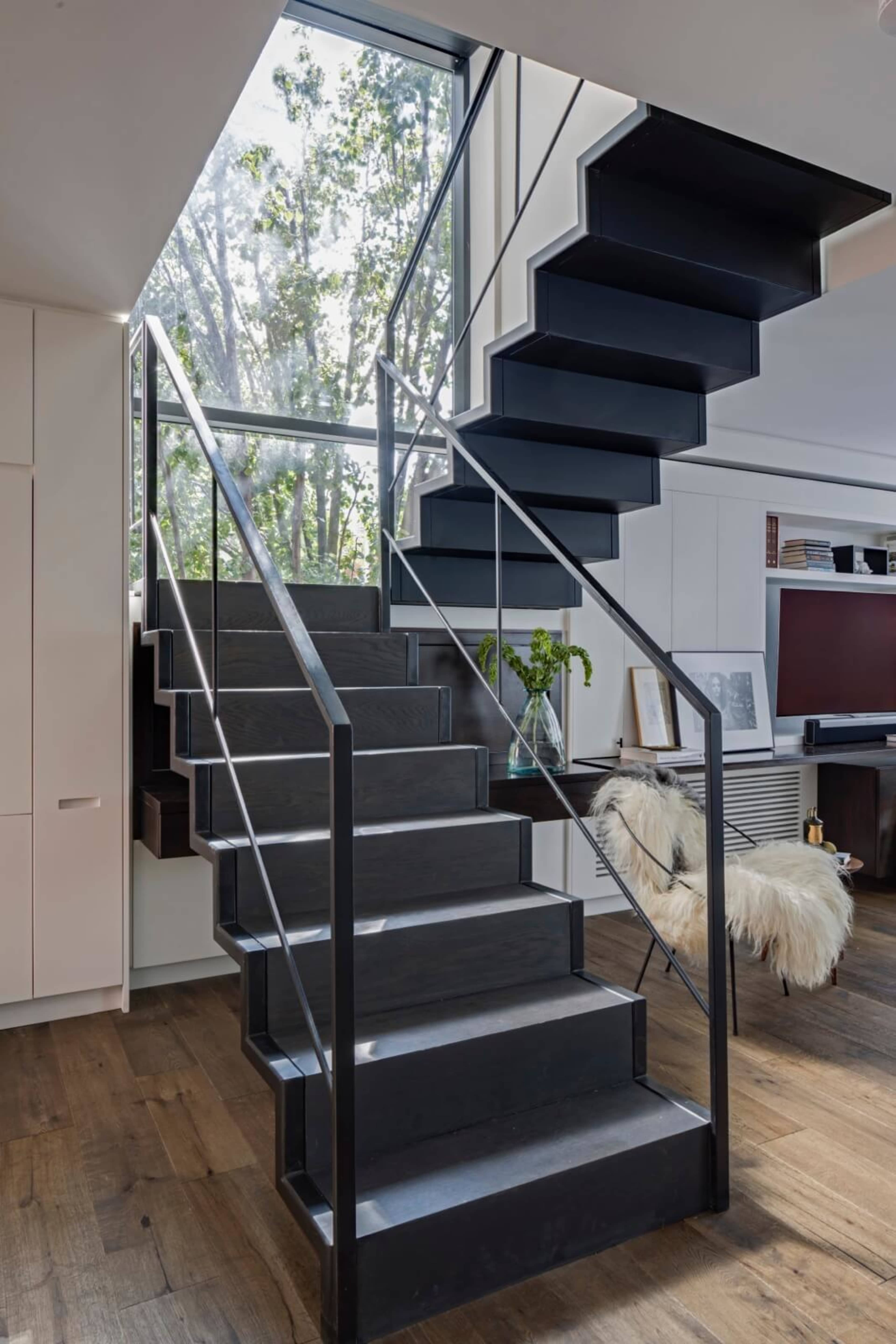 A modern staircase with dark wooden steps and metal railings leads to a glass-walled upper level illuminated by natural light.
