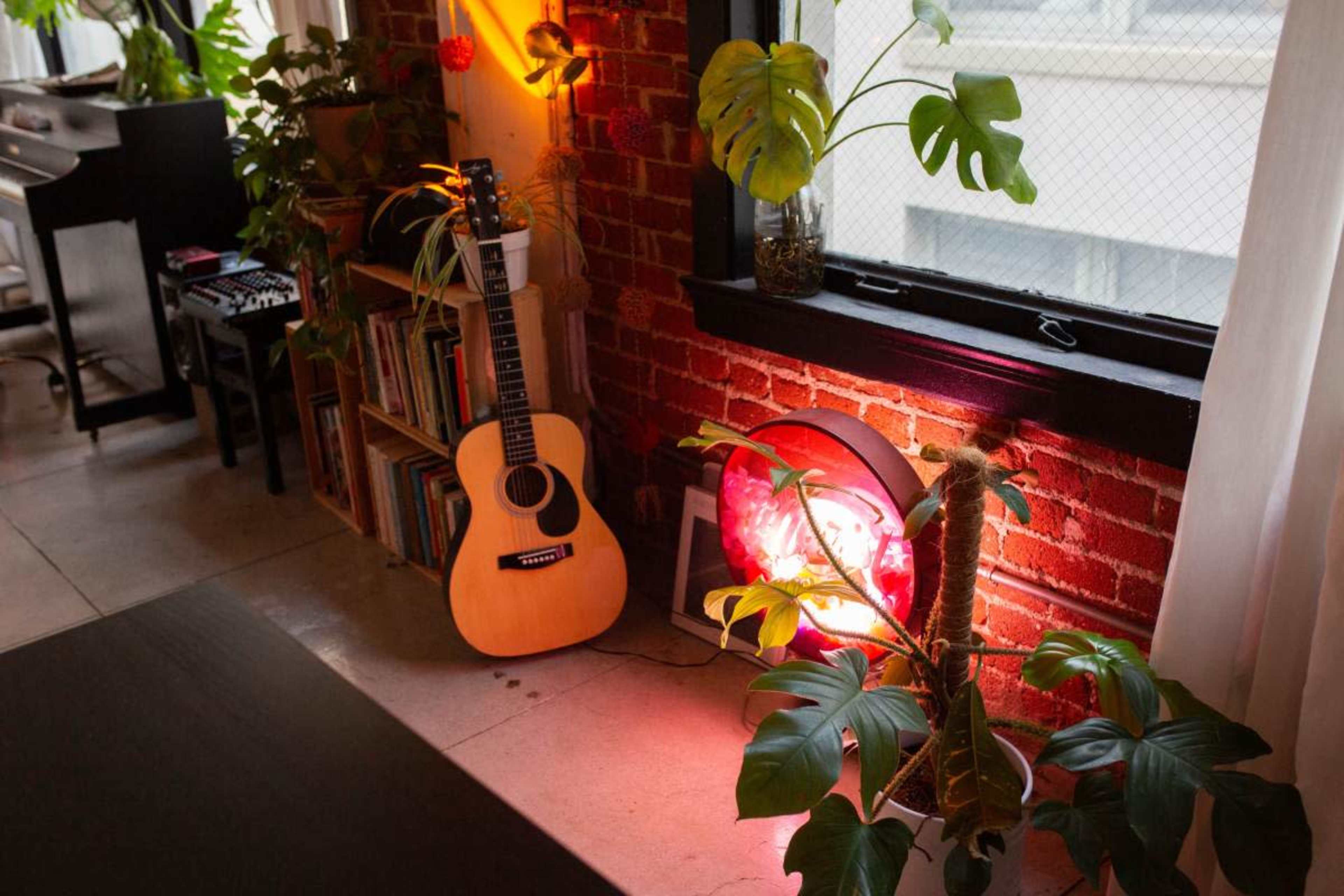 A wooden acoustic guitar leans against a brick wall next to a large red circular light and several potted plants.