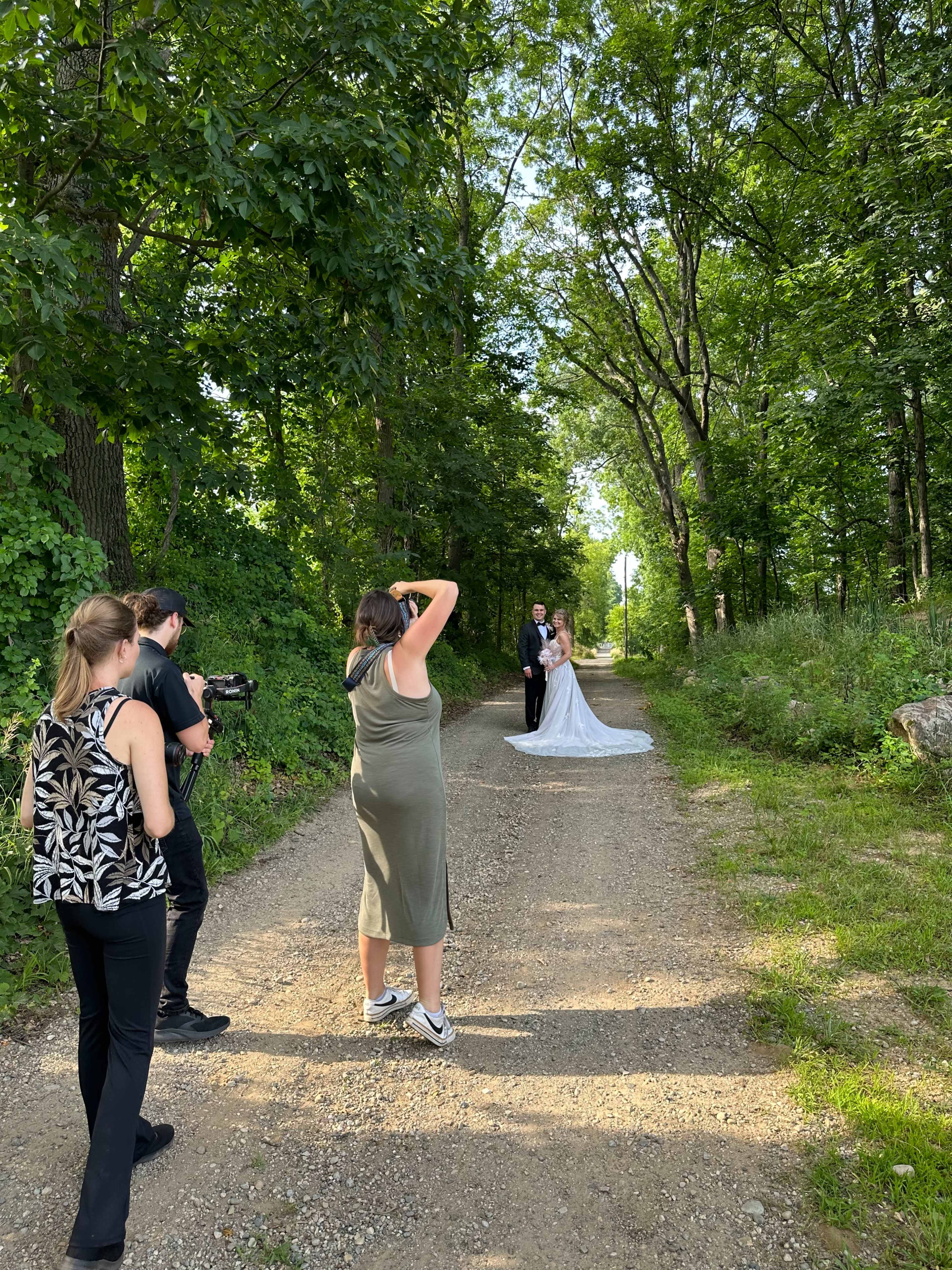A bride and groom stand on a dirt path surrounded by trees while a photographer captures their portrait.