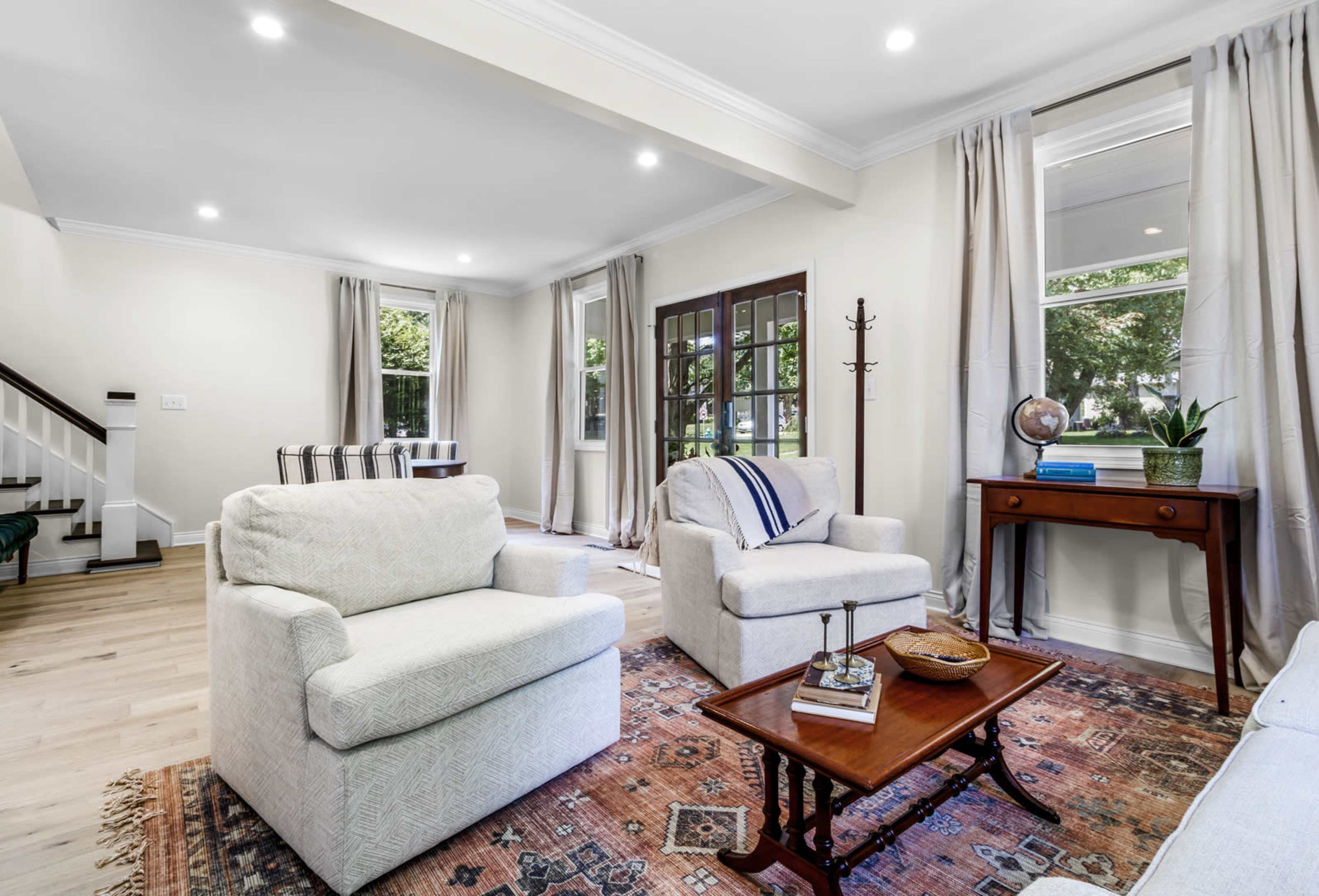 A bright living room with two light-colored armchairs, a wooden table, and large windows covered with curtains.