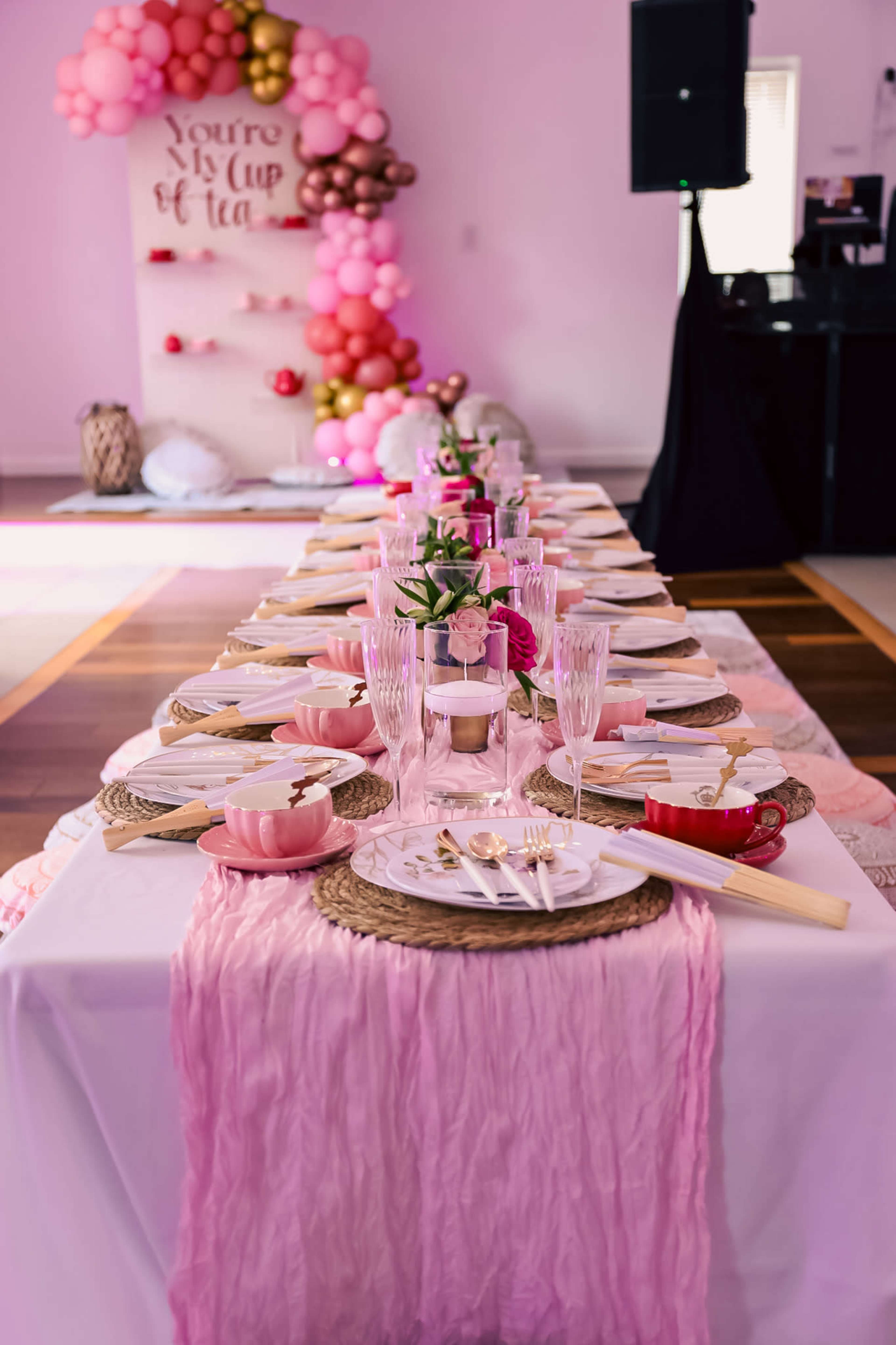 A long dining table is elegantly set with pink decorations, place settings, and a decorative backdrop featuring balloons.