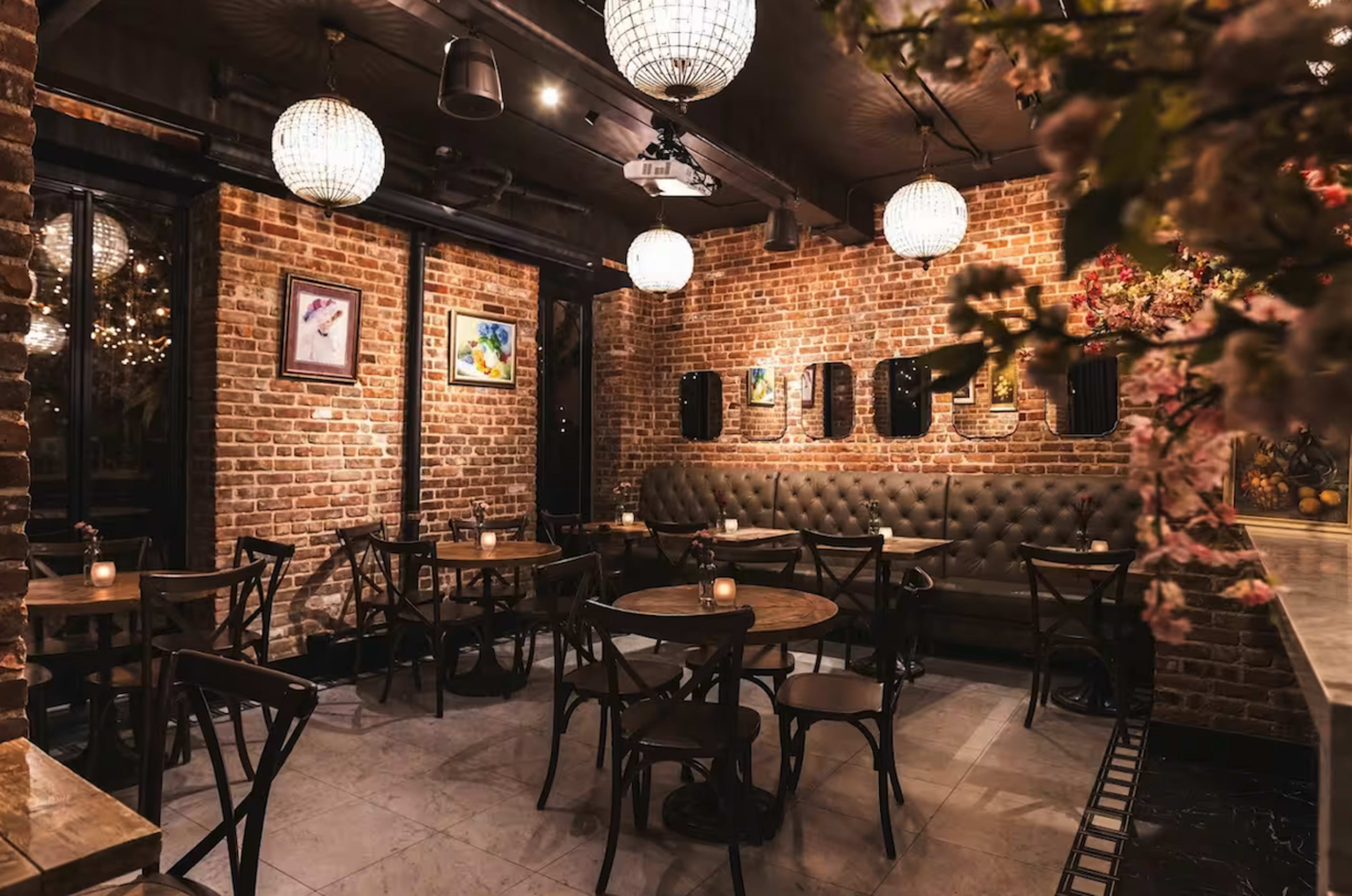 The image shows a dimly lit restaurant featuring exposed brick walls, round light fixtures, and wooden tables arranged around a cushioned seating area.