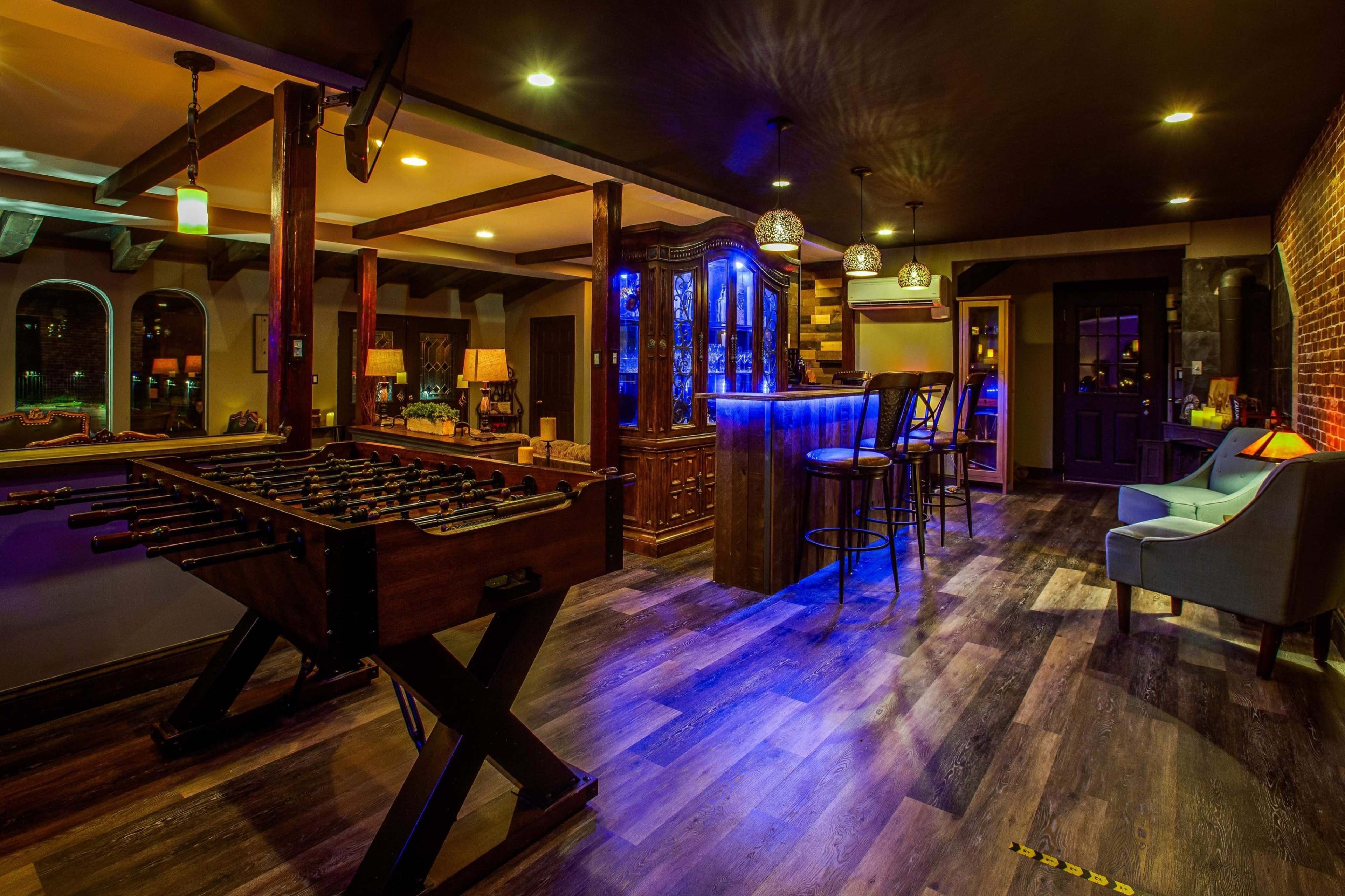 A dimly lit game room featuring a foosball table, a bar with blue lighting, and seating area with a couch and chairs.