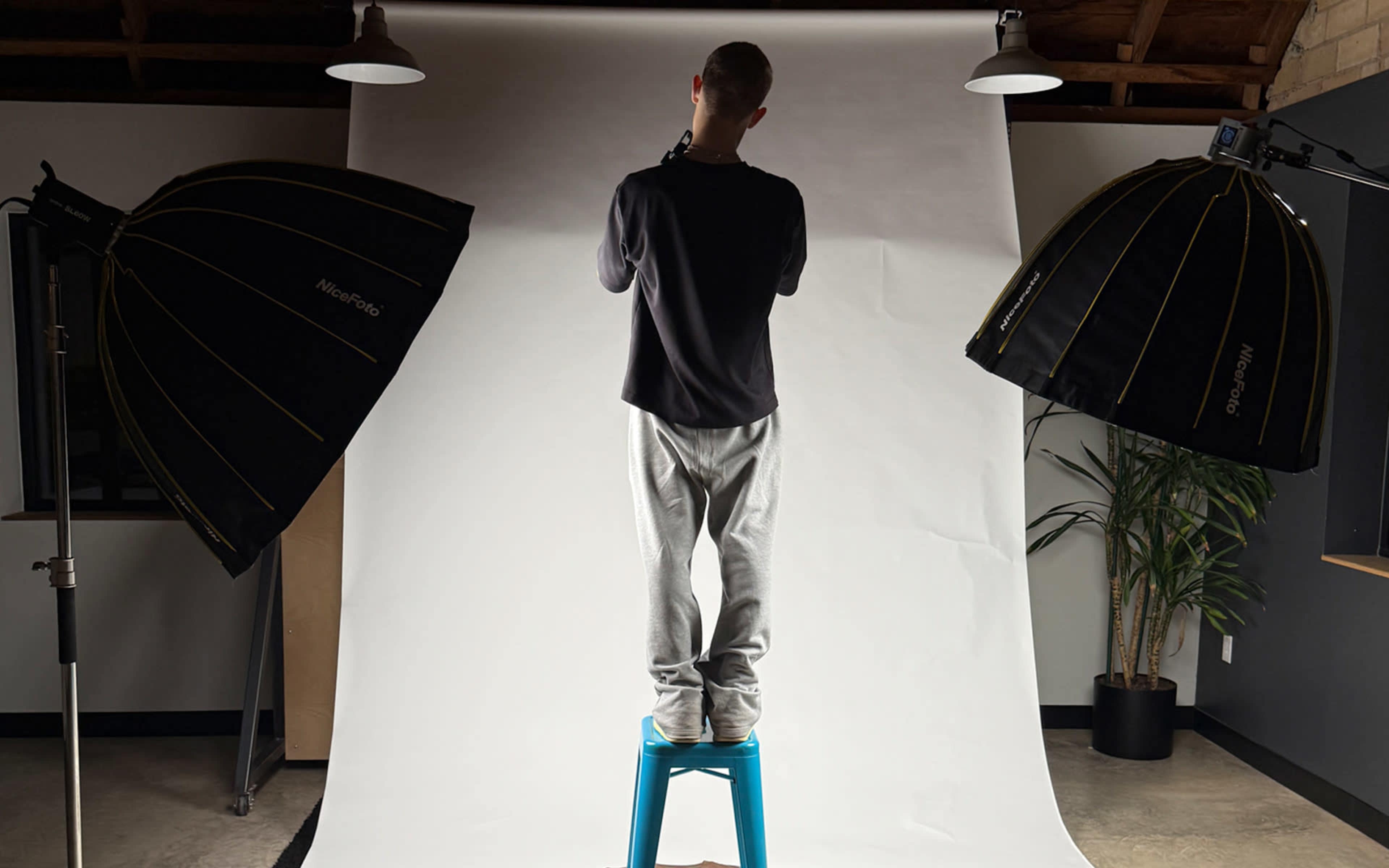 A person stands on a blue stool in front of a white backdrop, flanked by two large studio light modifiers.