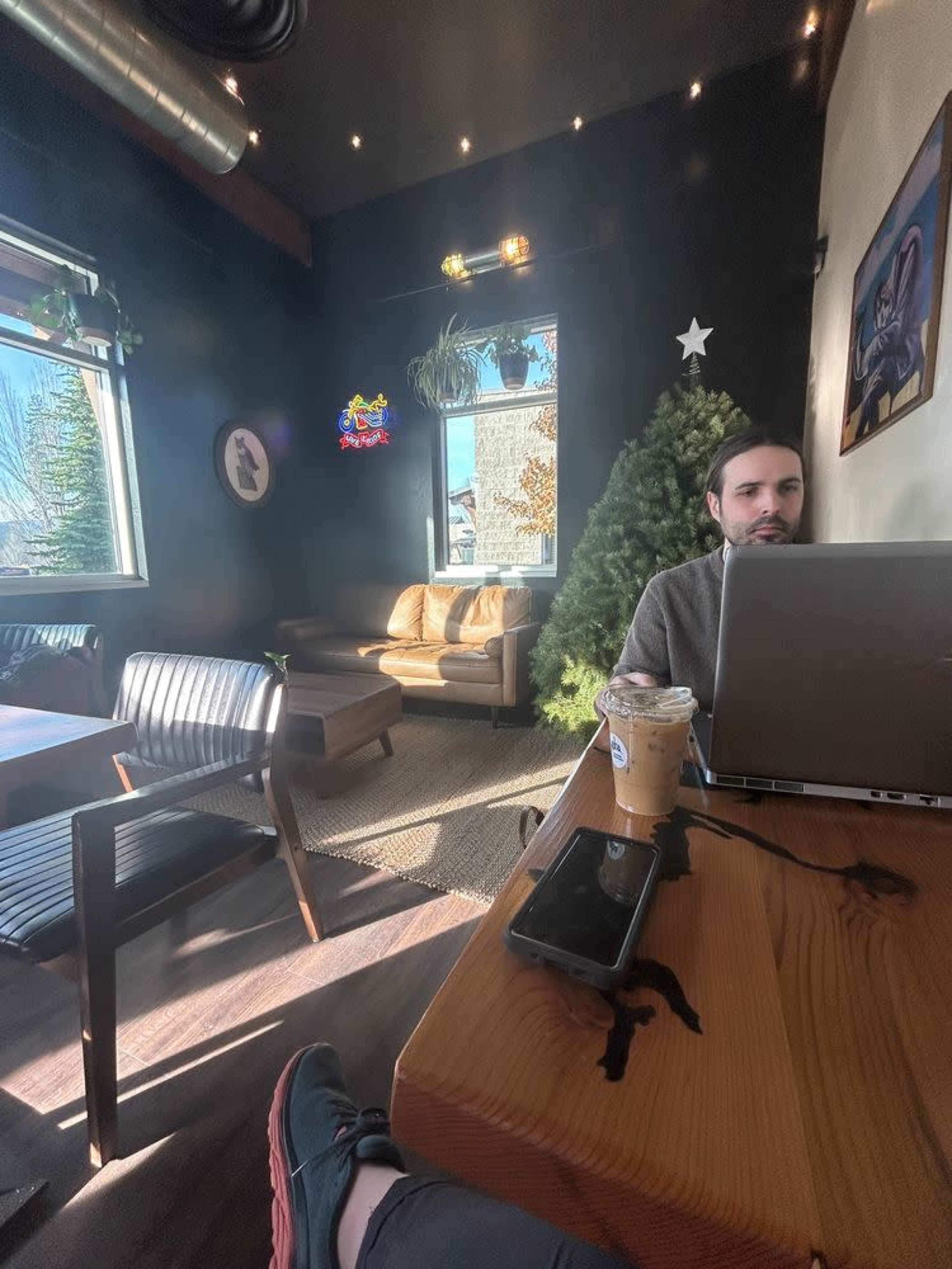 A person sits at a wooden table in a coffee shop, working on a laptop with an iced drink nearby, while a Christmas tree decorates the corner.