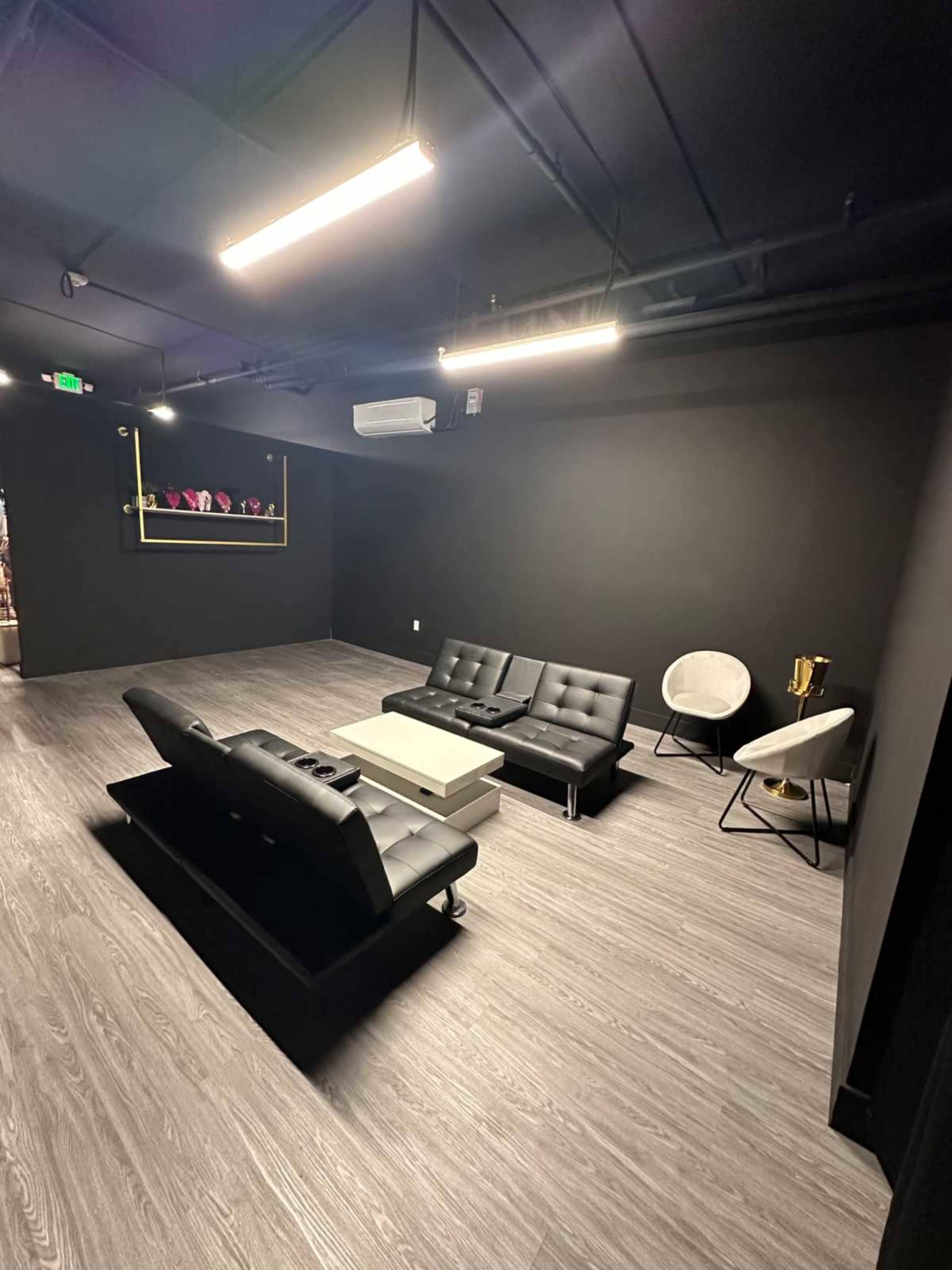 The image shows a modern lounge area with black walls, a sleek black sofa, a white coffee table, and a white chair, illuminated by bright overhead lights.