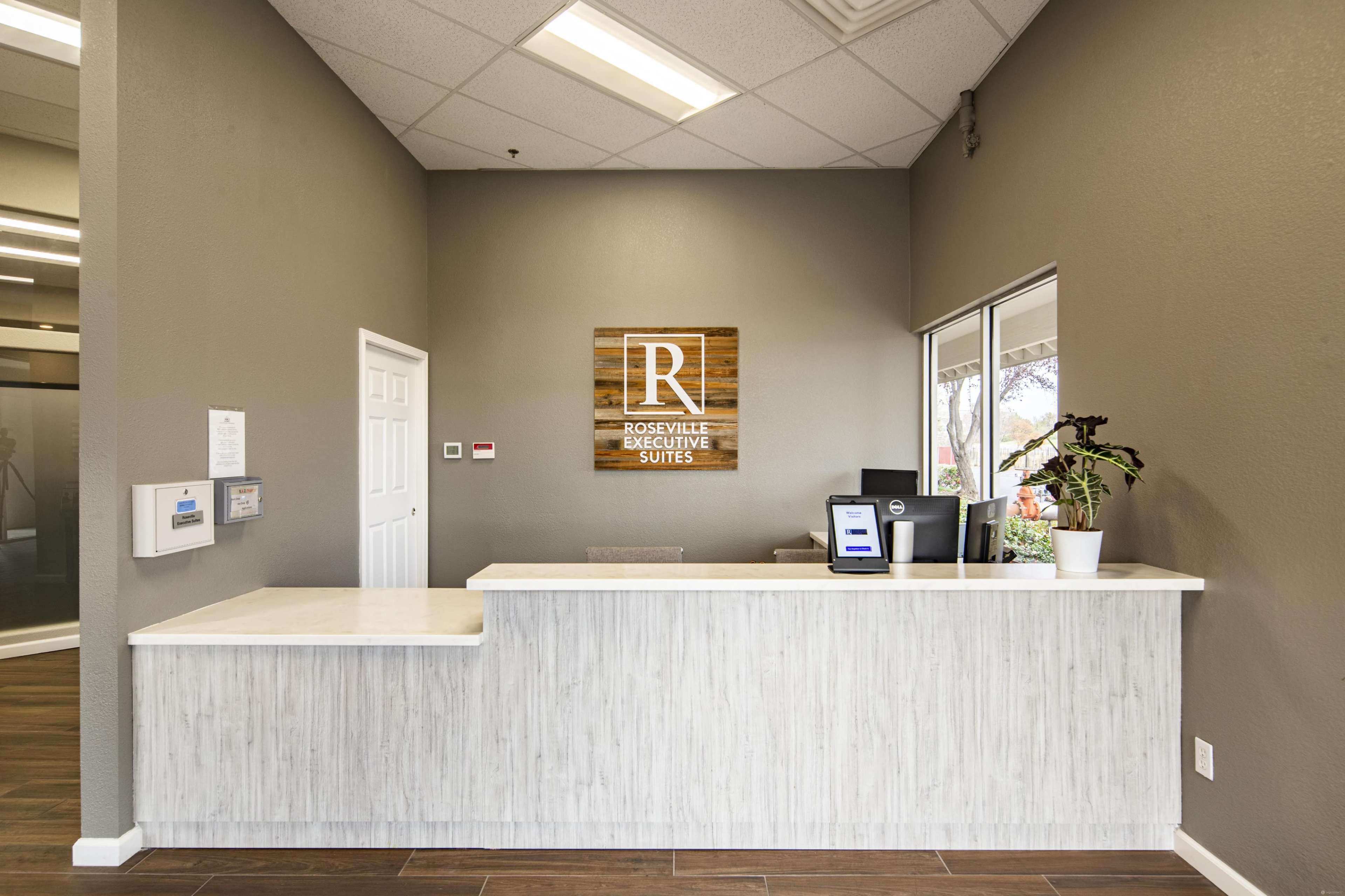The image shows a modern reception desk at Roseville Executive Suites, featuring a sign with the letter "R" and a digital display.