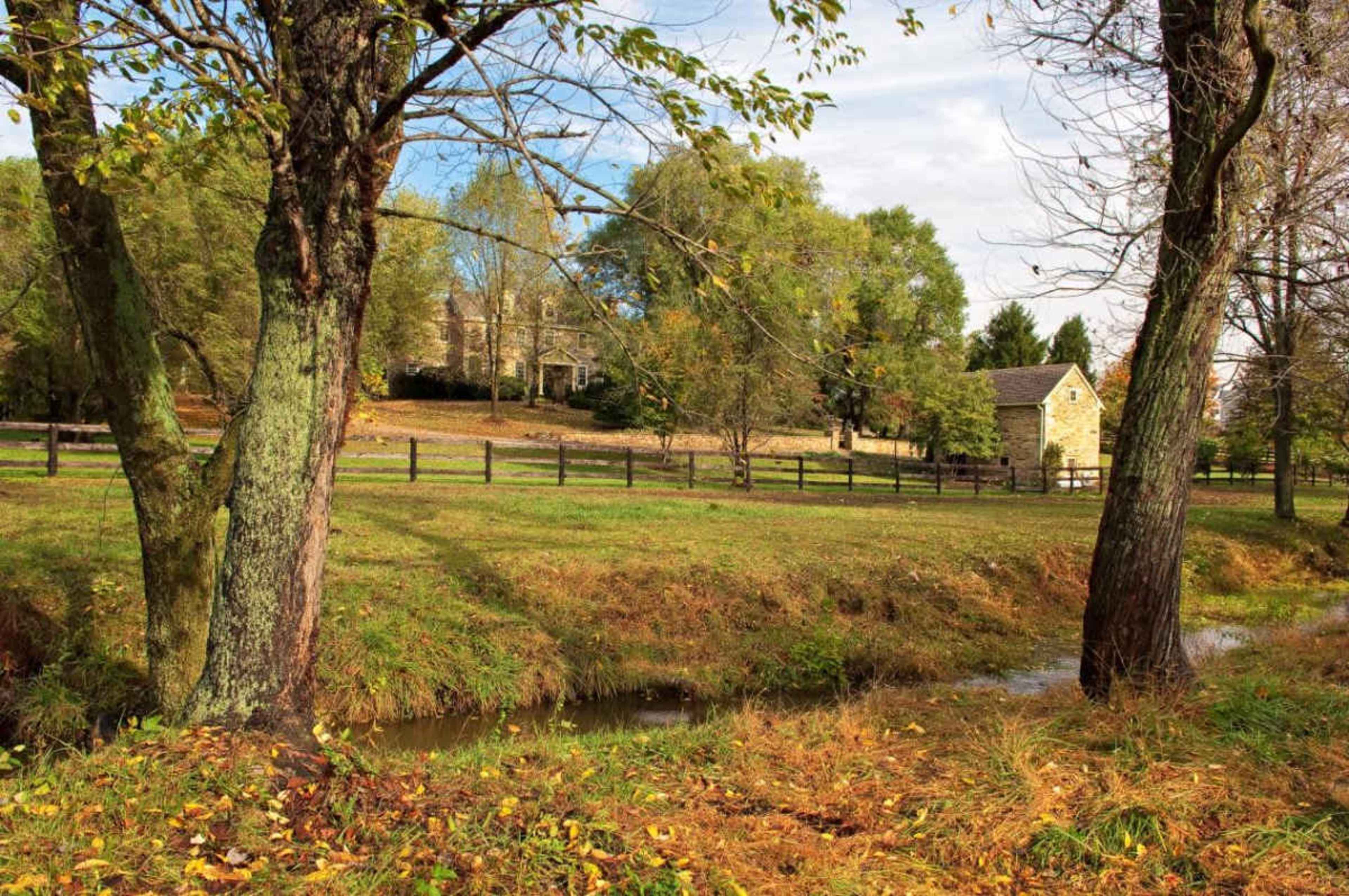 A shallow stream runs alongside a grassy area with trees, leading to a stone house in the background, surrounded by a wooden fence.