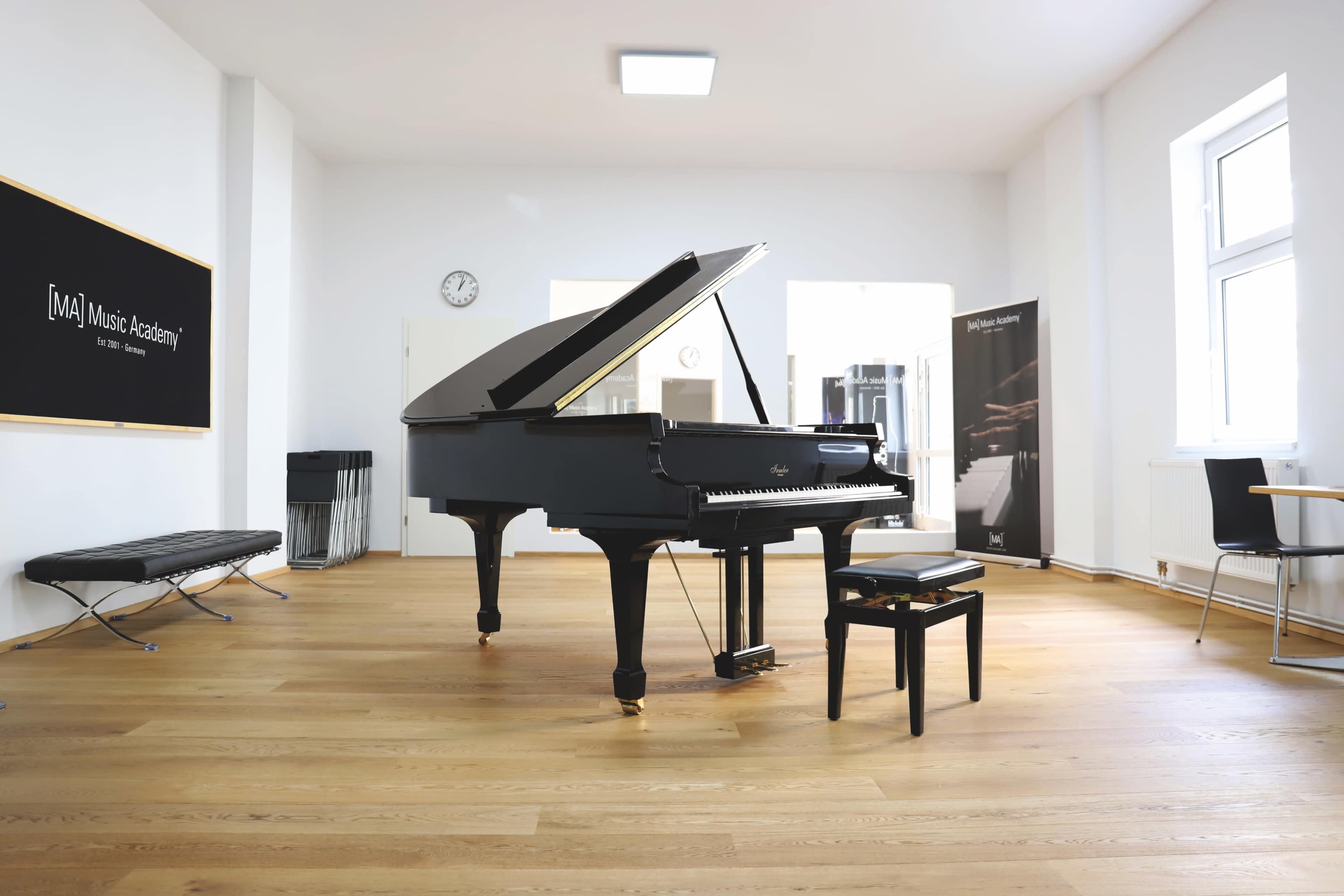 A grand piano is set in a spacious, well-lit music studio with wooden flooring and a black bench beside it.