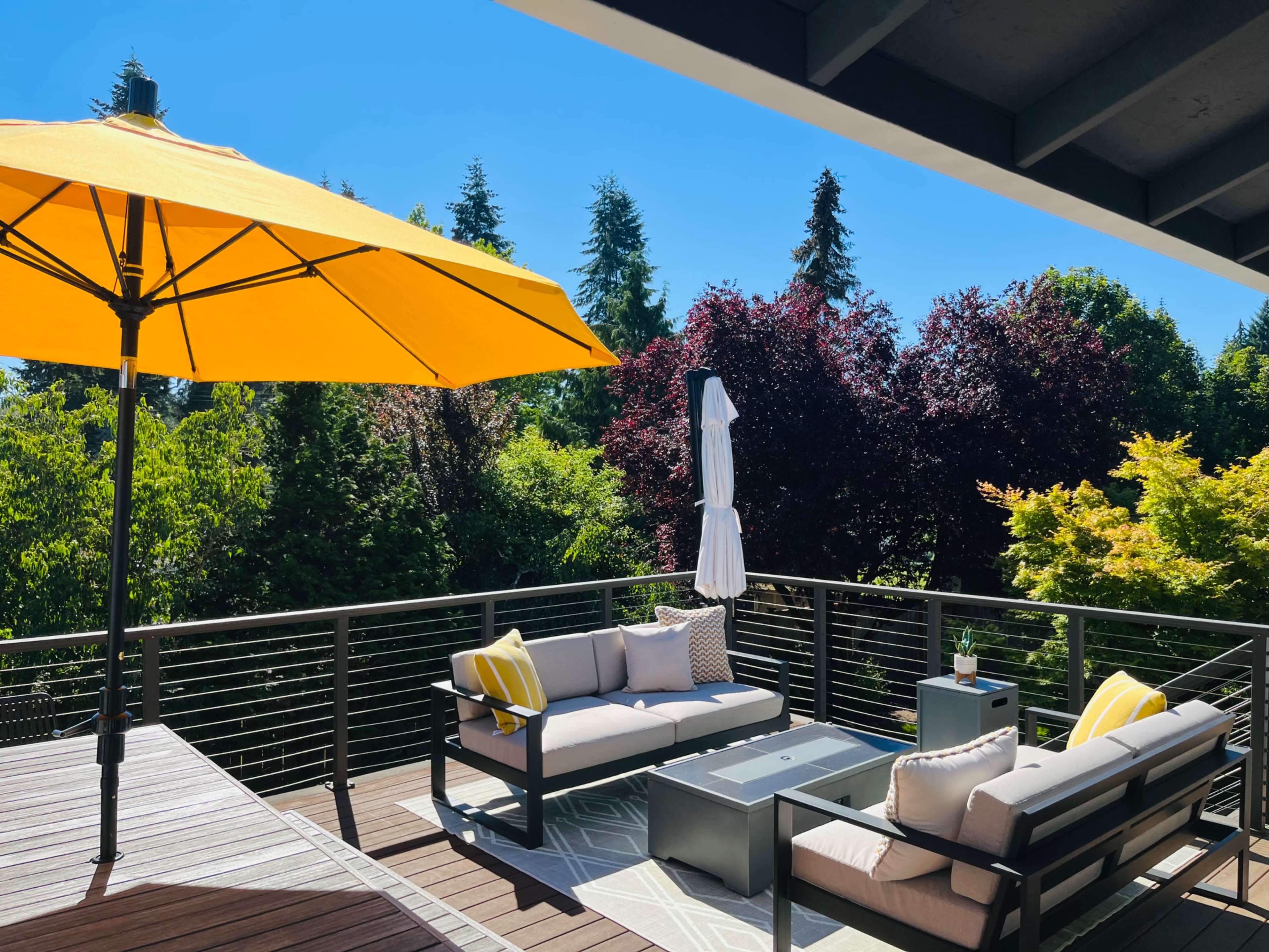A deck featuring a yellow umbrella, grey seating with cushions, and surrounding lush trees.