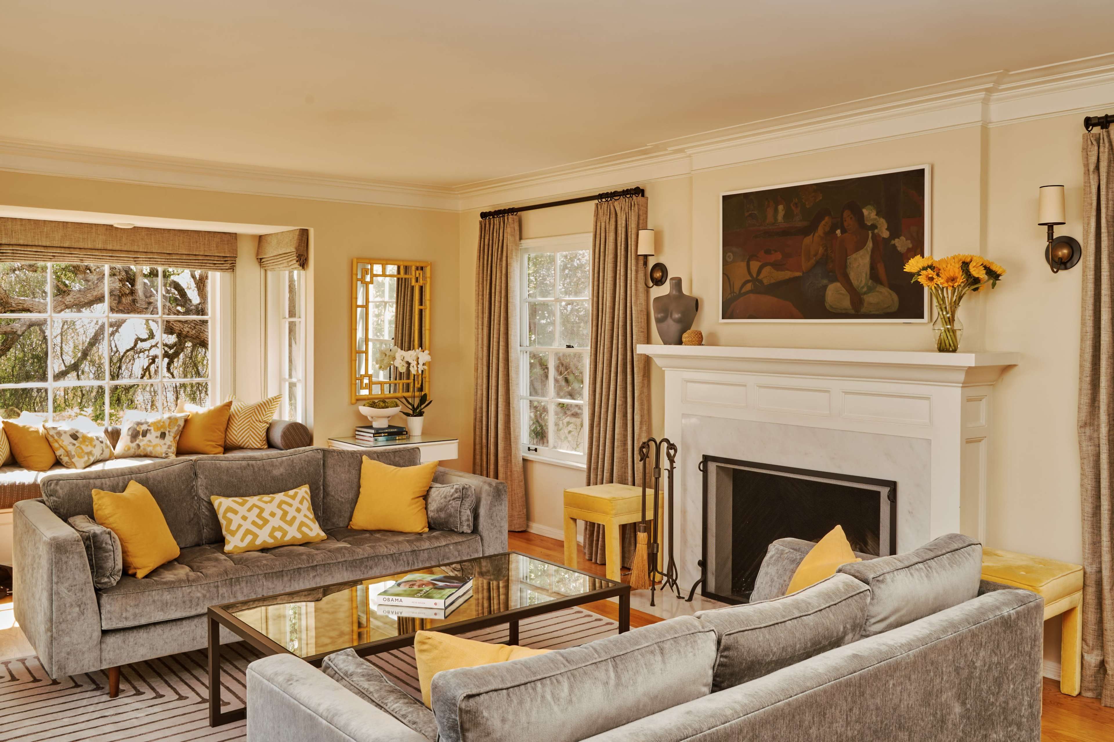 A bright living room features two gray sofas adorned with yellow pillows, a glass coffee table, and a fireplace with a modern painting above it, illuminated by natural light from large windows.