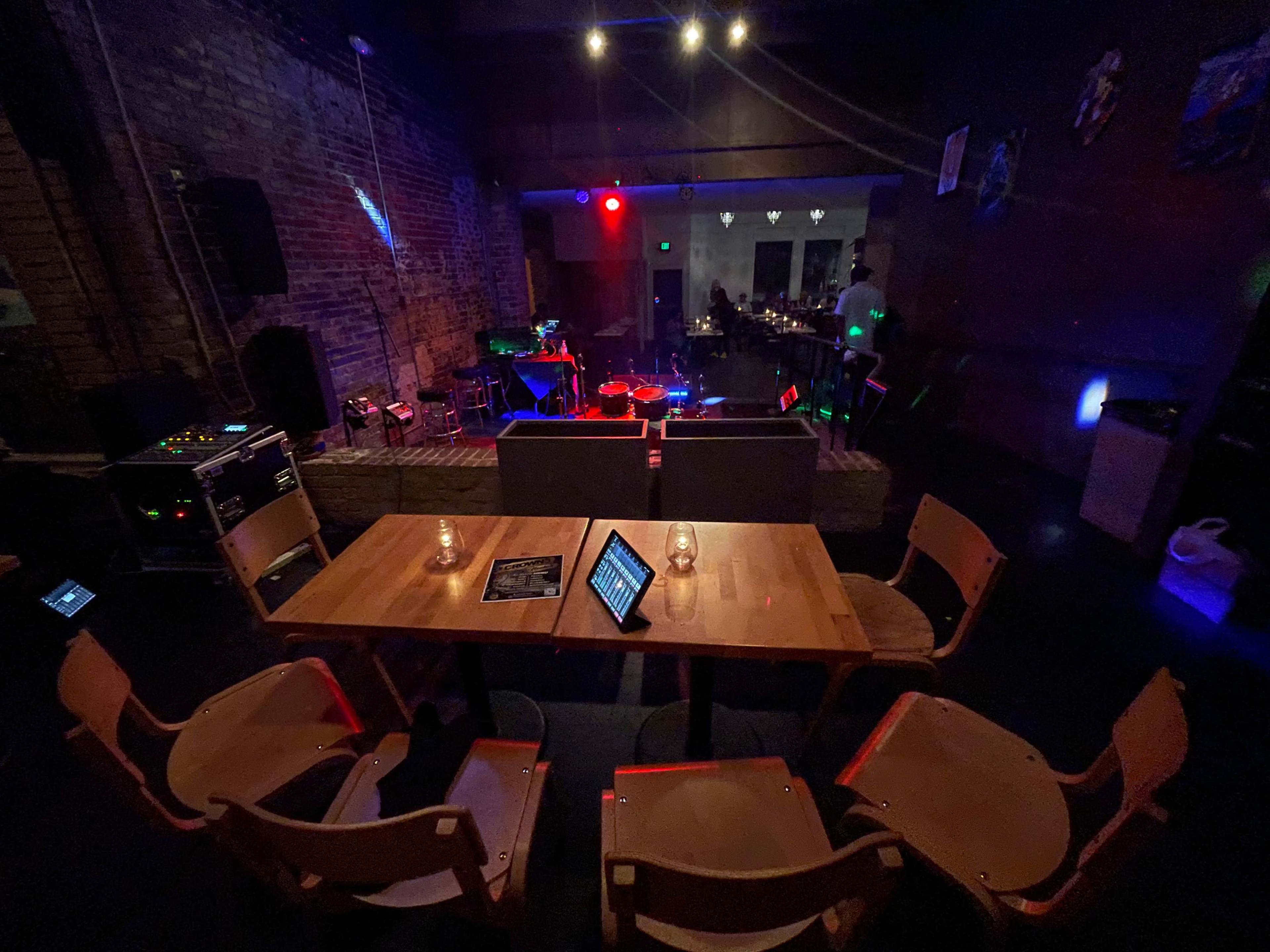 An empty restaurant or bar setting with wooden tables and chairs, soft lighting, and a stage area in the background.
