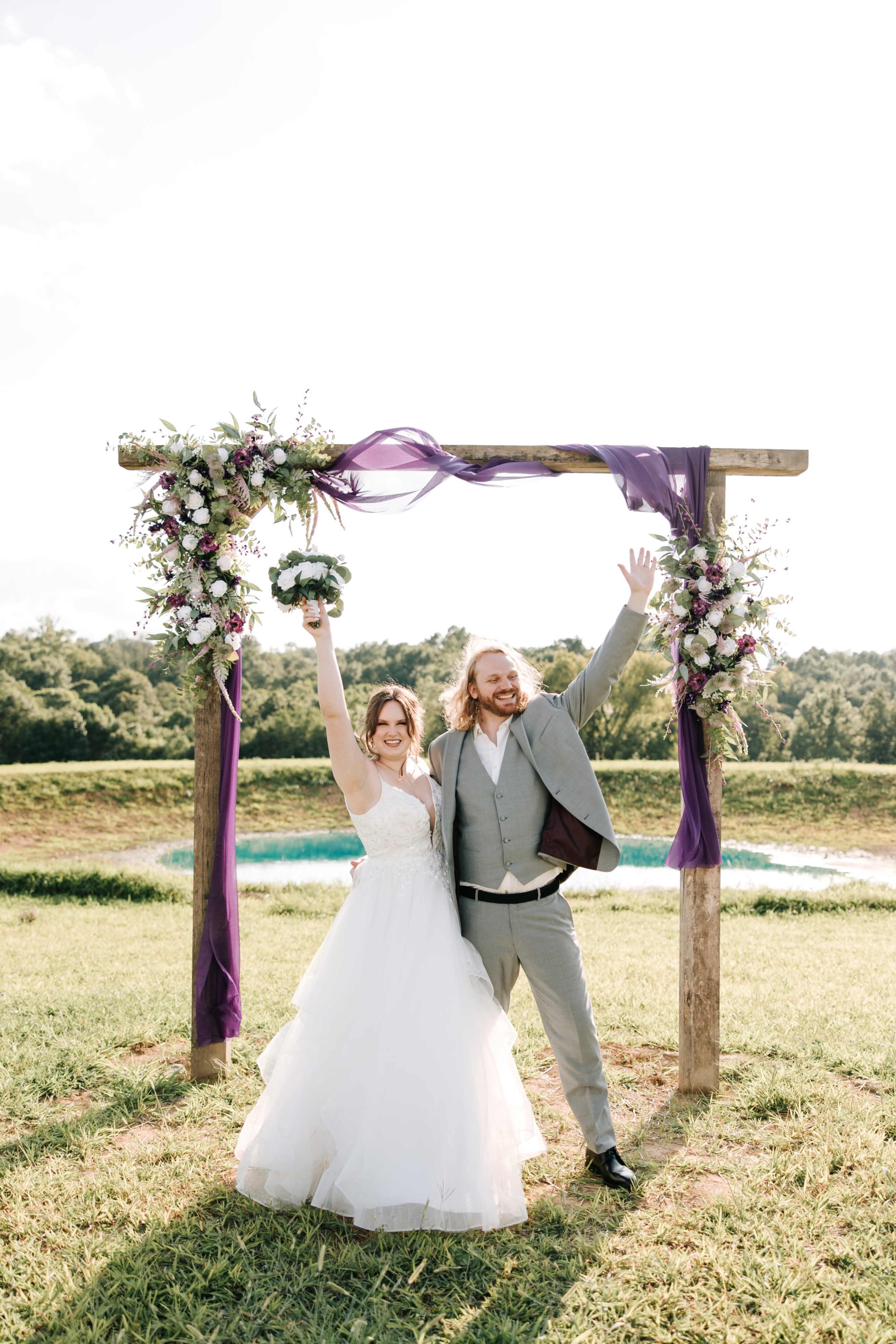 A bride and groom stand joyfully in front of a flower-adorned archway at an outdoor wedding.