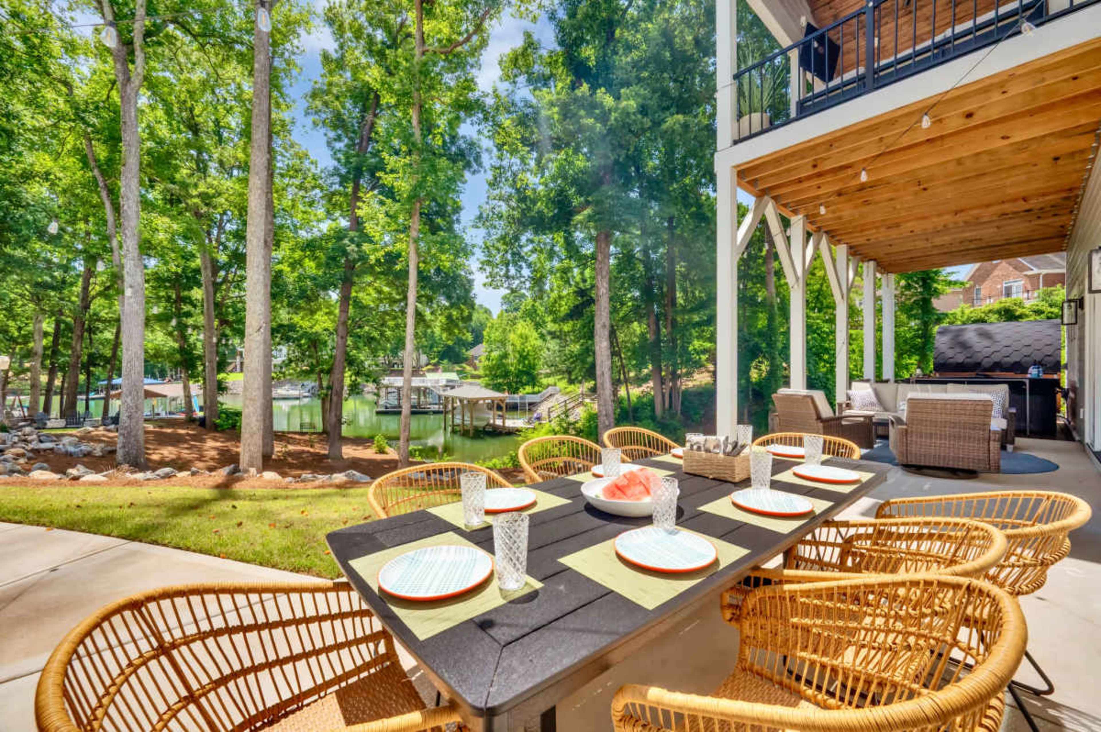 A patio dining area with a large table and woven chairs overlooks a serene waterfront surrounded by trees.