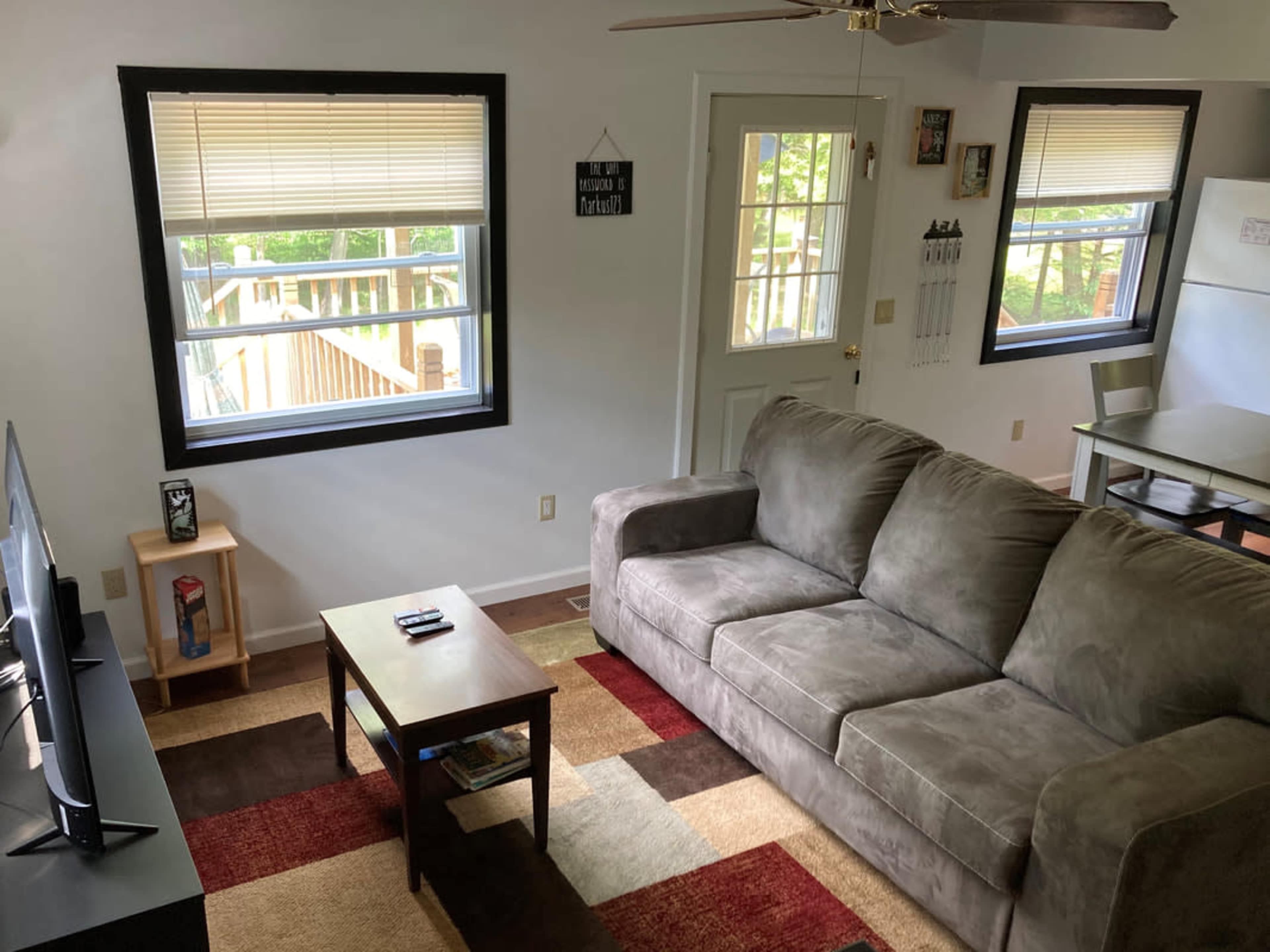 A living room with a beige couch, a coffee table, a television on a stand, and two windows facing a wooden deck outside.