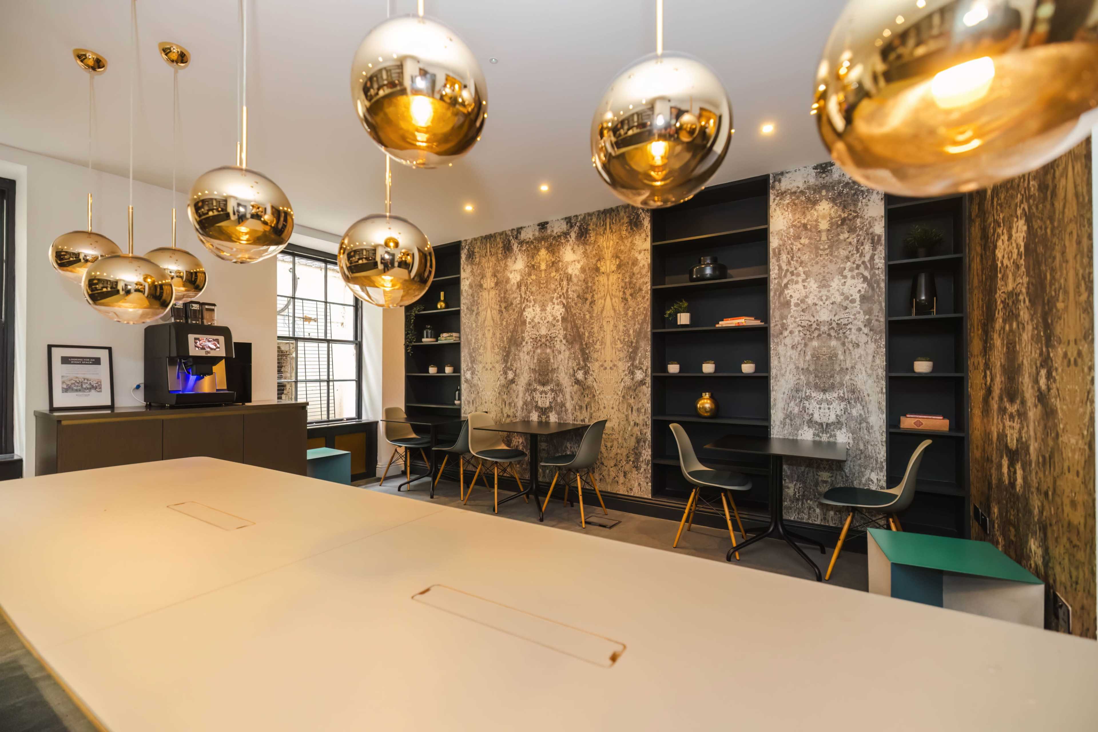The image shows a modern office space featuring a long table in the foreground, golden pendant lights hanging above, and a textured wall with shelves and seating areas in the background.
