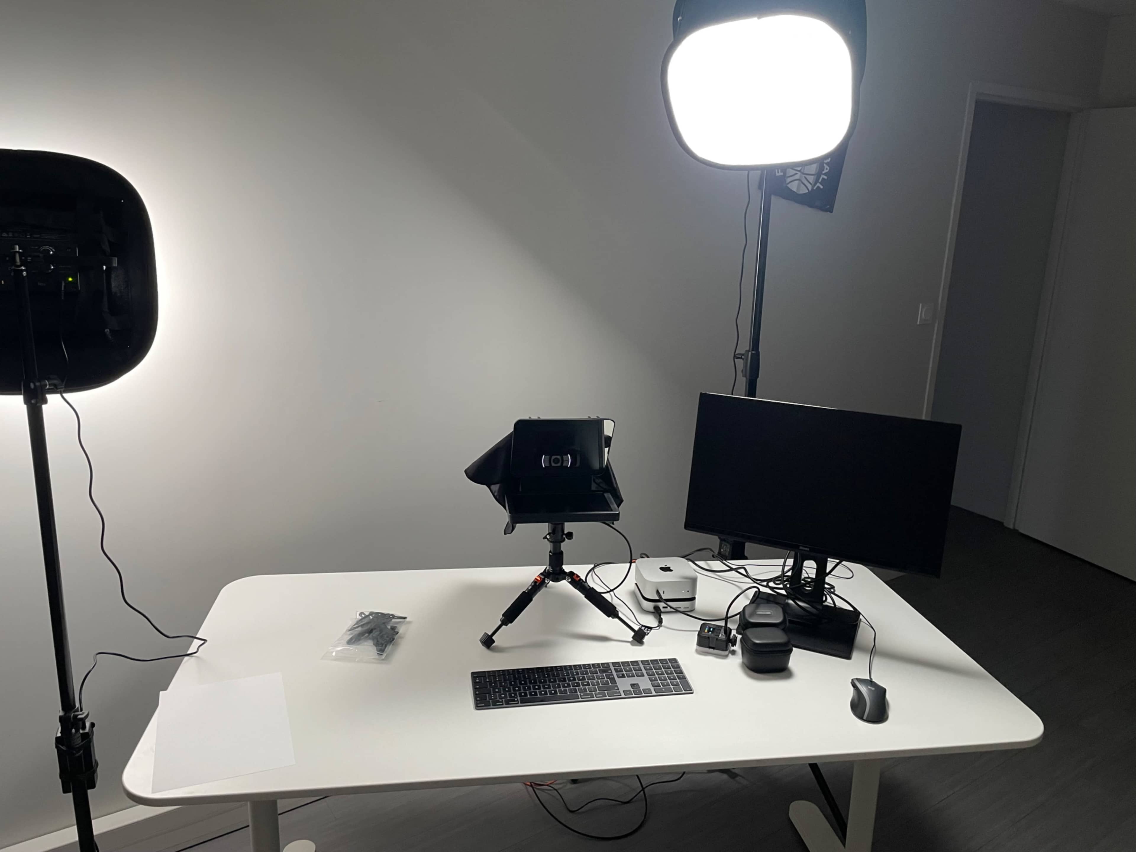 The image shows a workspace setup featuring a desk with a monitor, a keyboard, a mouse, and a camera on a tripod next to two light sources.