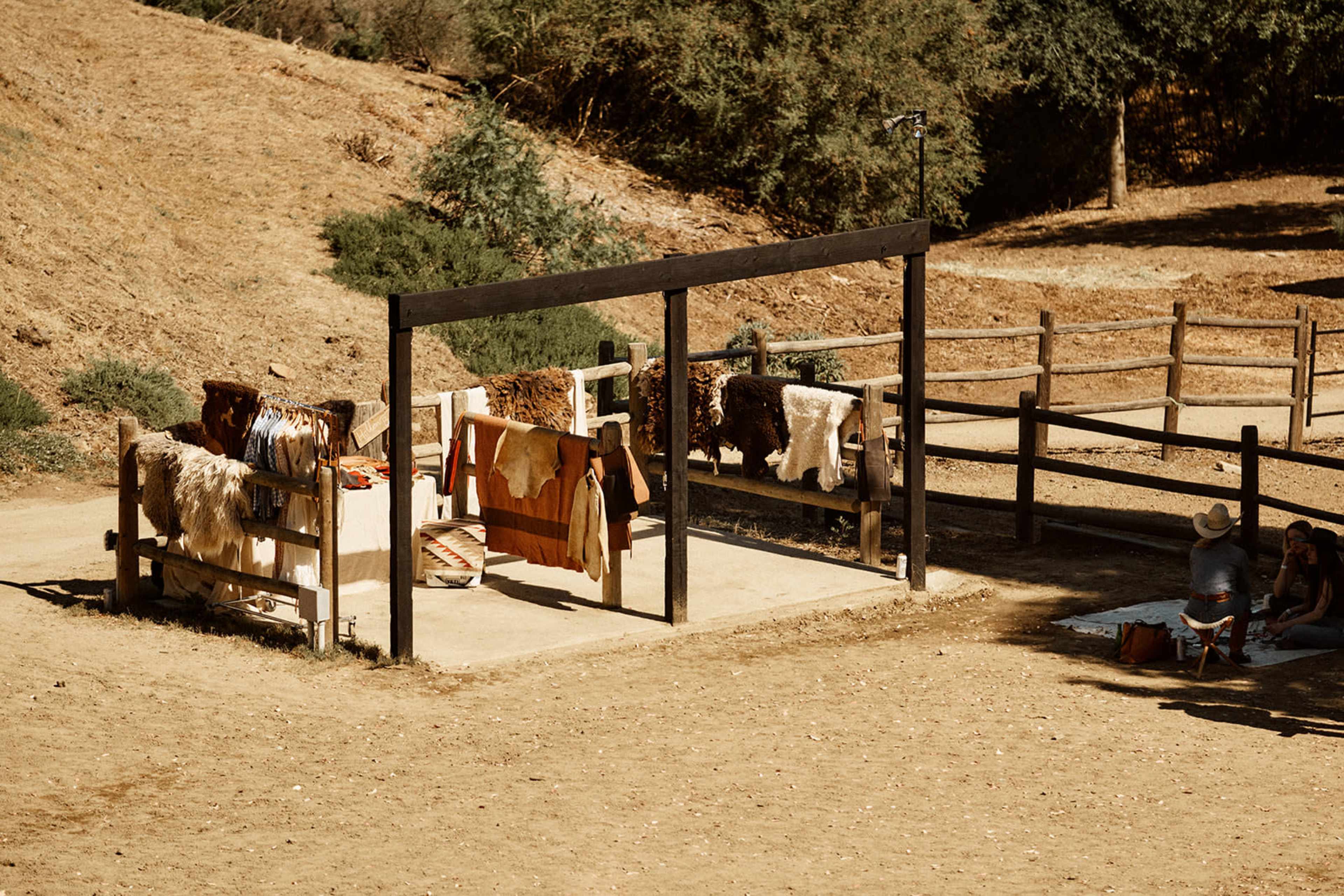 The image shows a rustic outdoor area with several animal hides displayed on wooden frames near a dirt path and a fenced enclosure.