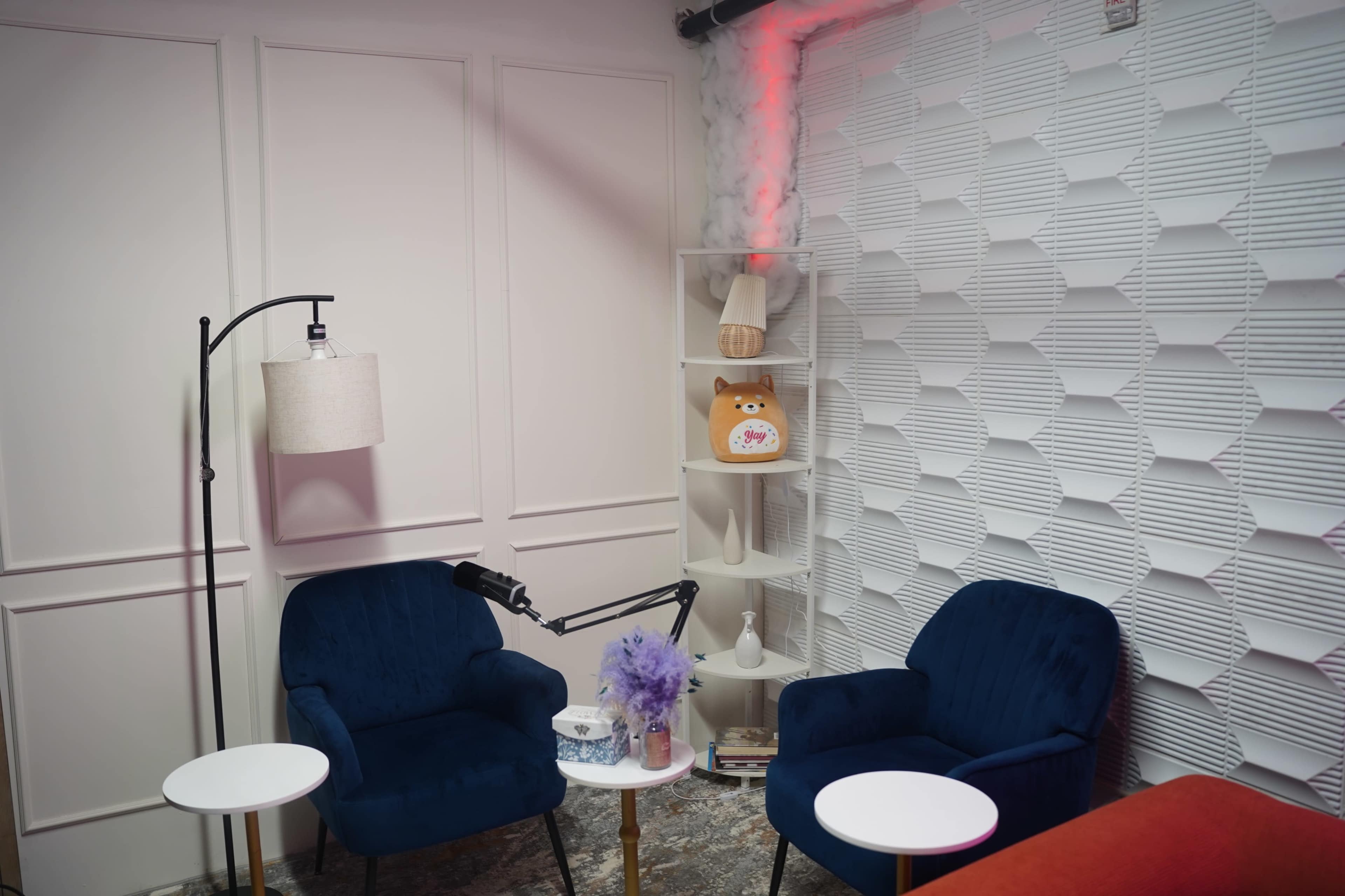 The image shows a cozy corner of a room featuring two blue chairs, a small white table, a lamp, and a shelf with decorative items against a textured wall.