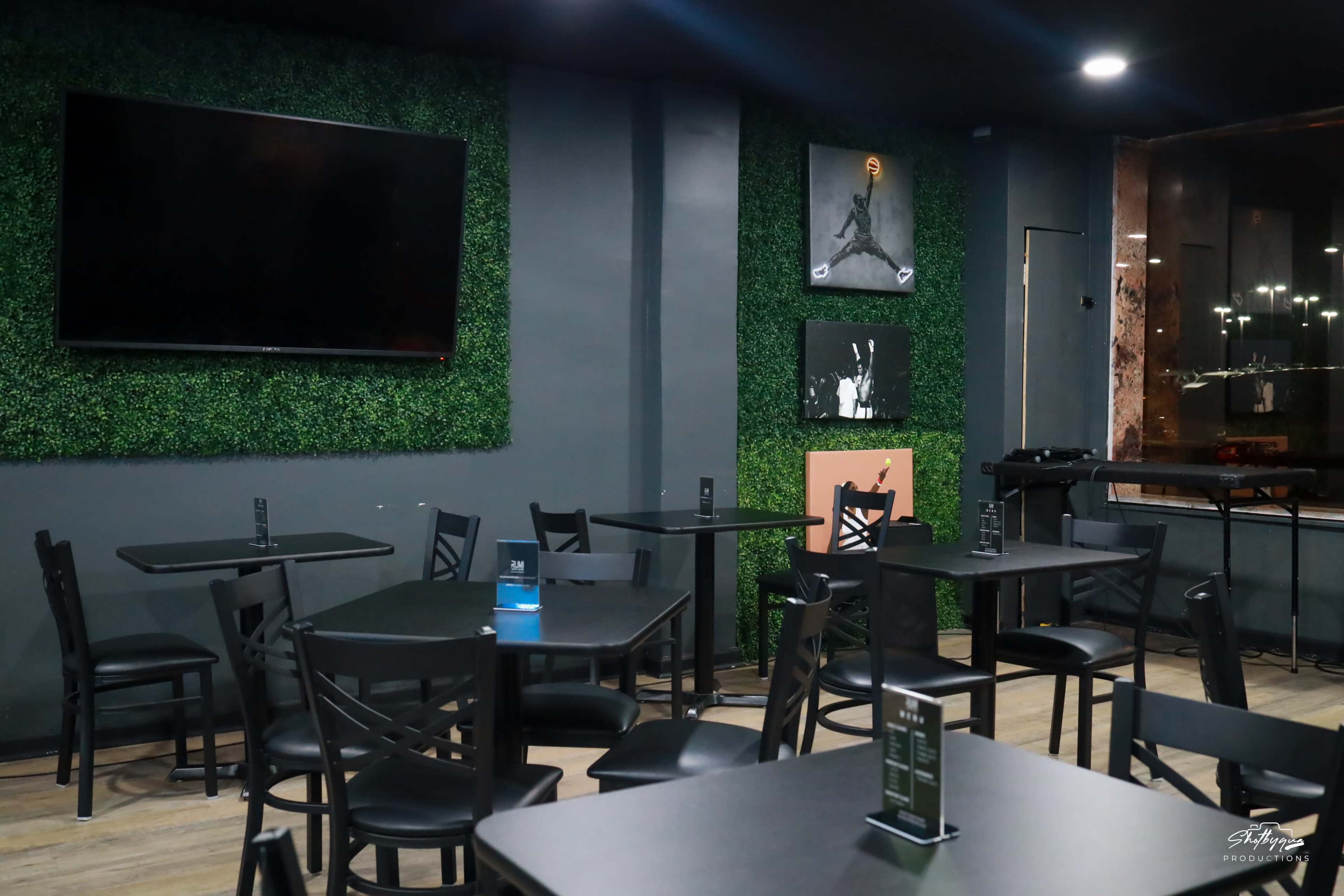 A dimly lit restaurant interior featuring black tables and chairs against a green wall with artificial greenery, complemented by a wall-mounted television and framed artwork.