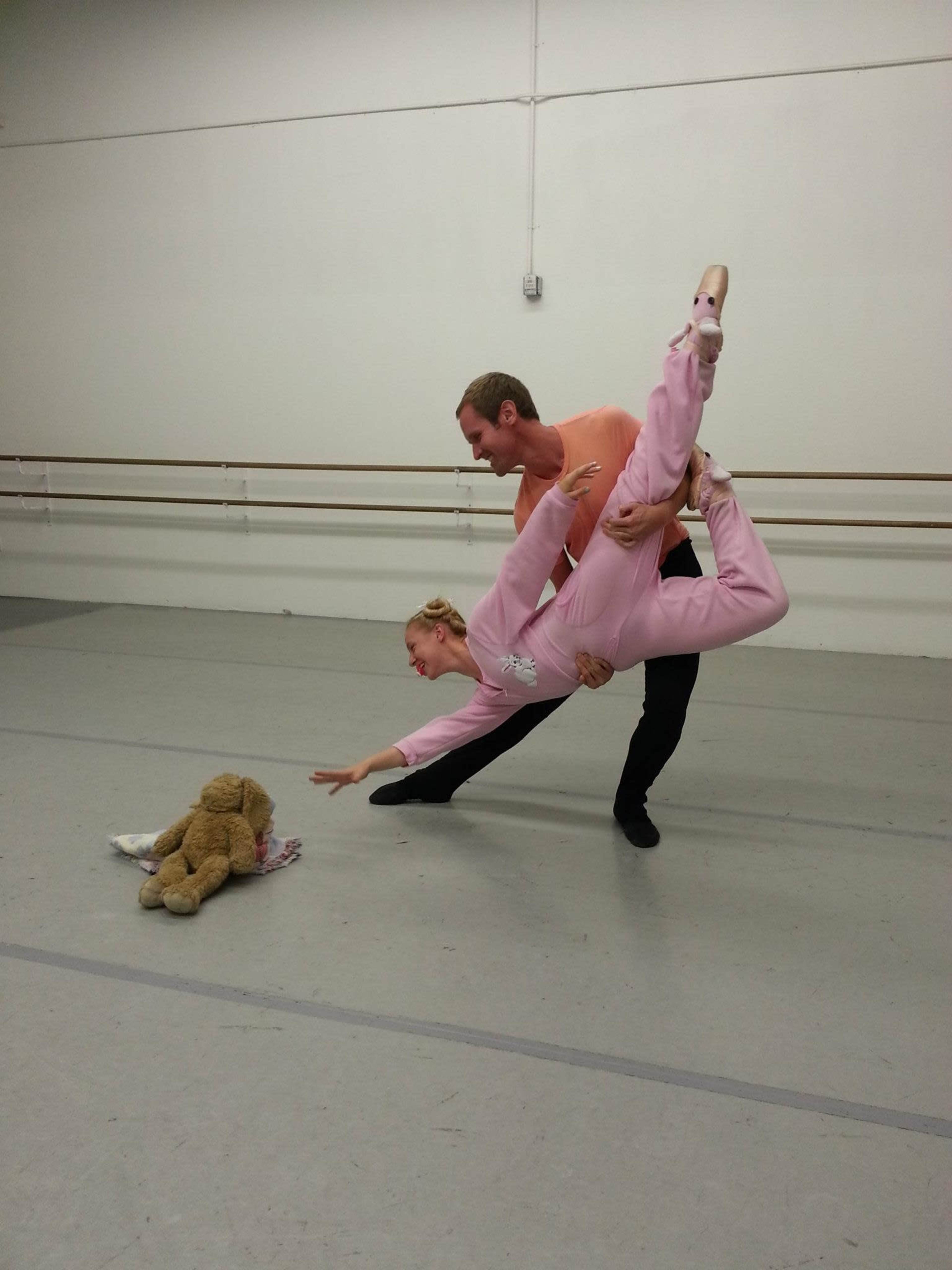 A dancer in a pink outfit is being lifted by a partner in an orange shirt, while a teddy bear lies on the floor nearby.