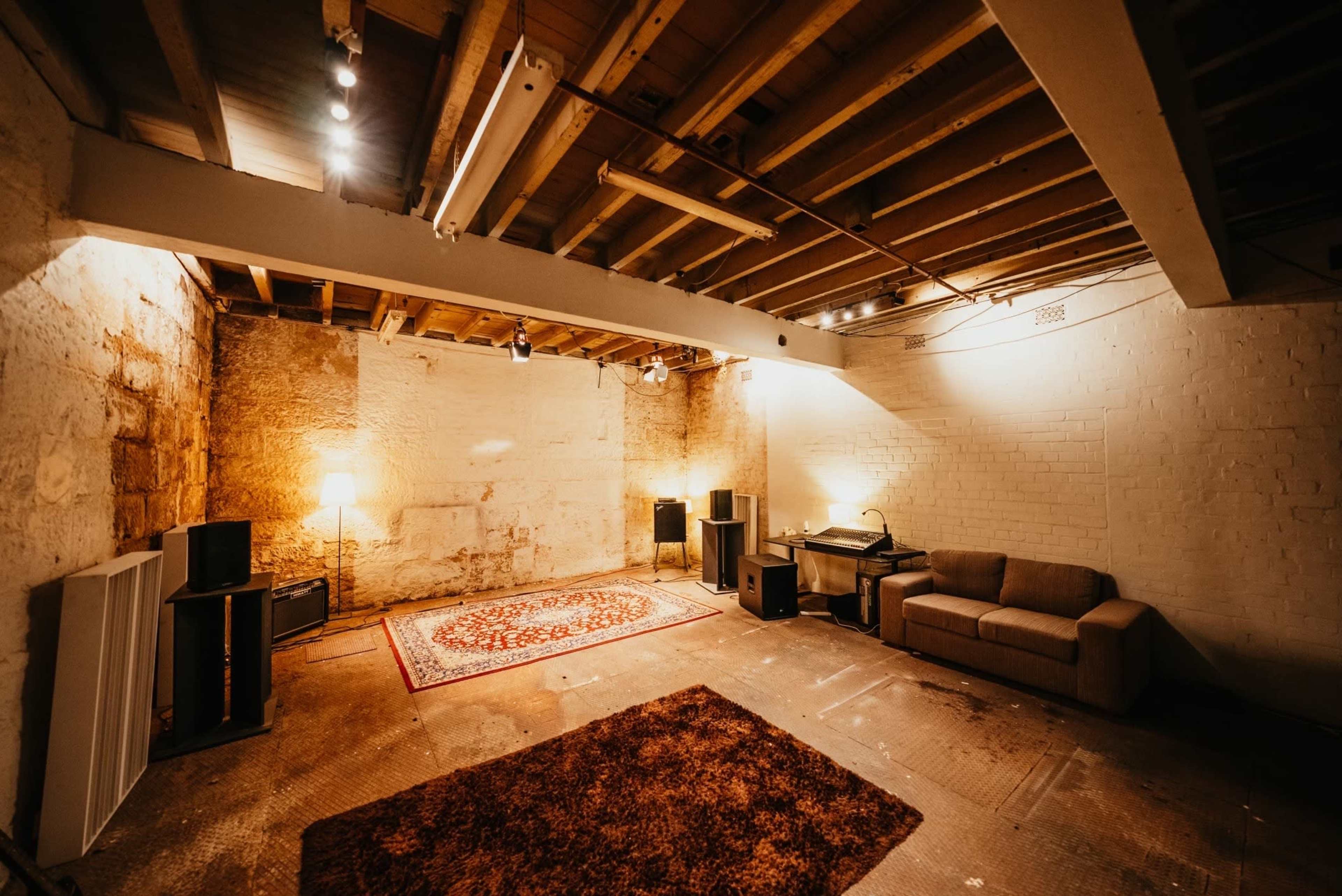 A spacious, partially finished basement with exposed wooden beams, a patterned rug on the floor, a couch, speakers, and soft lighting coming from a lamp.