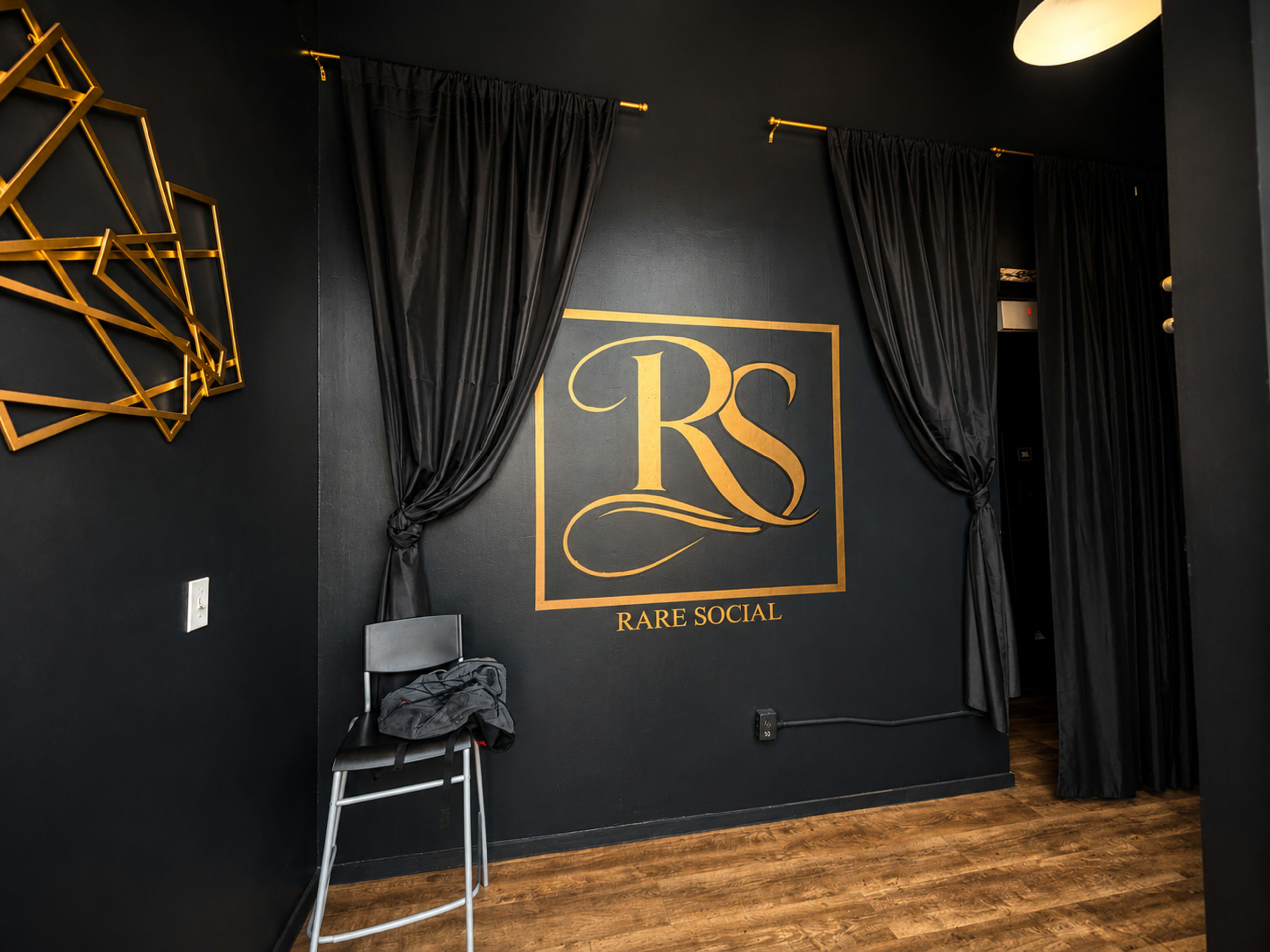 The image shows a darkly painted interior wall featuring a gold logo for "Rare Social" and two black curtains, with a metal chair positioned nearby.