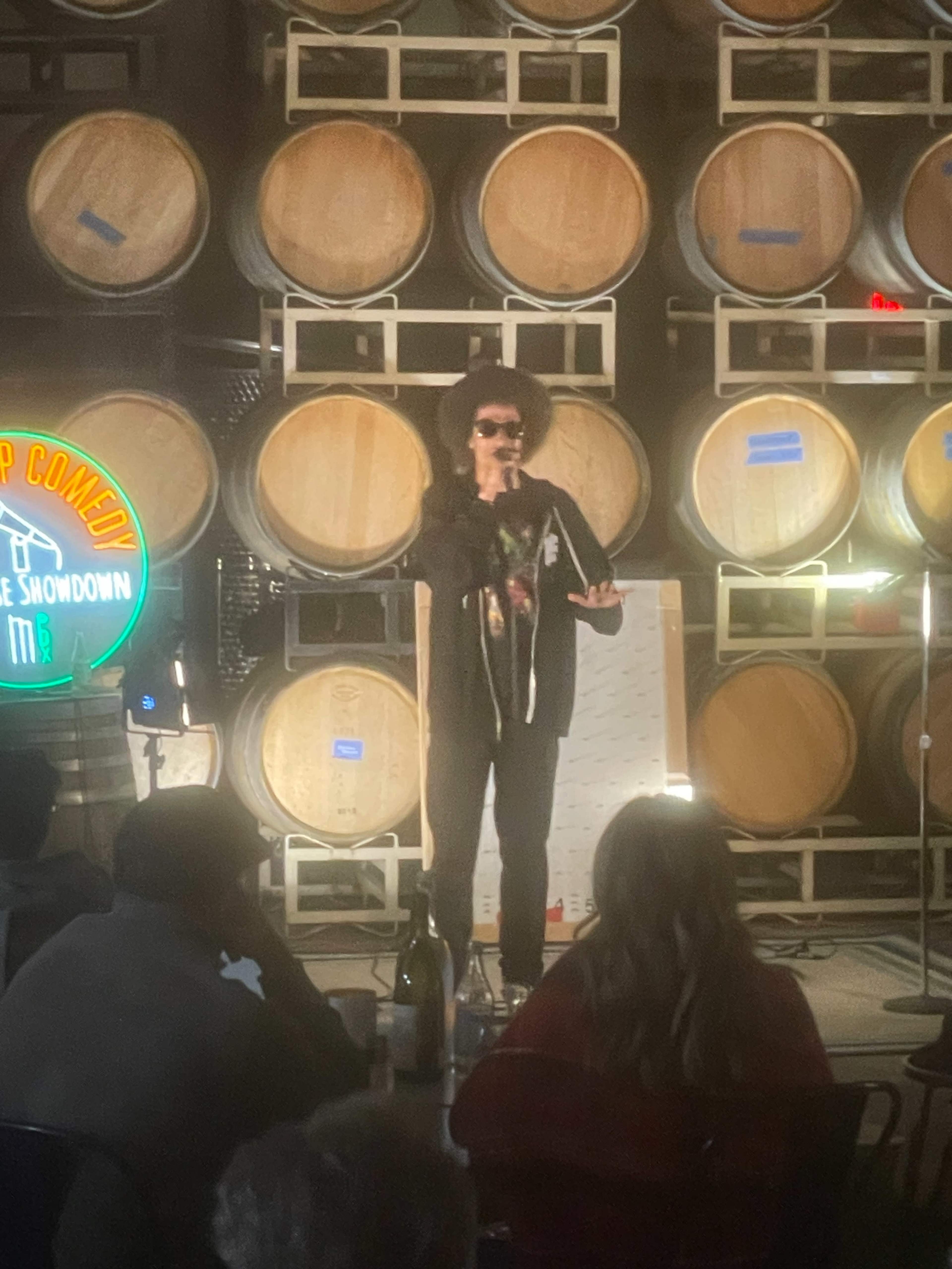 A performer stands on stage with a microphone in front of a backdrop of stacked wooden wine barrels.