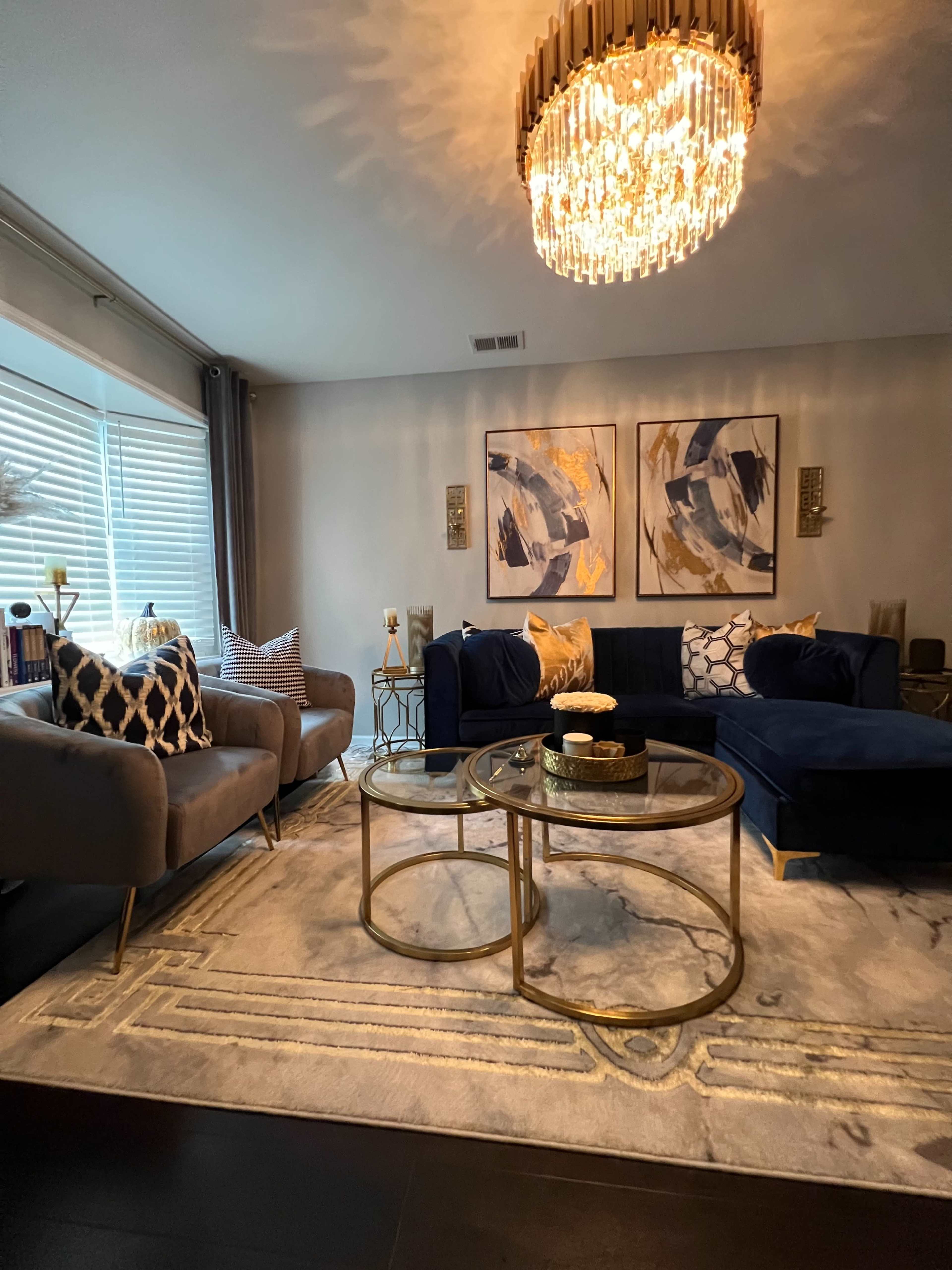 The living room features a blue sectional sofa, two gray armchairs, a pair of gold coffee tables on a patterned rug, and decorative wall art beneath a chandelier.