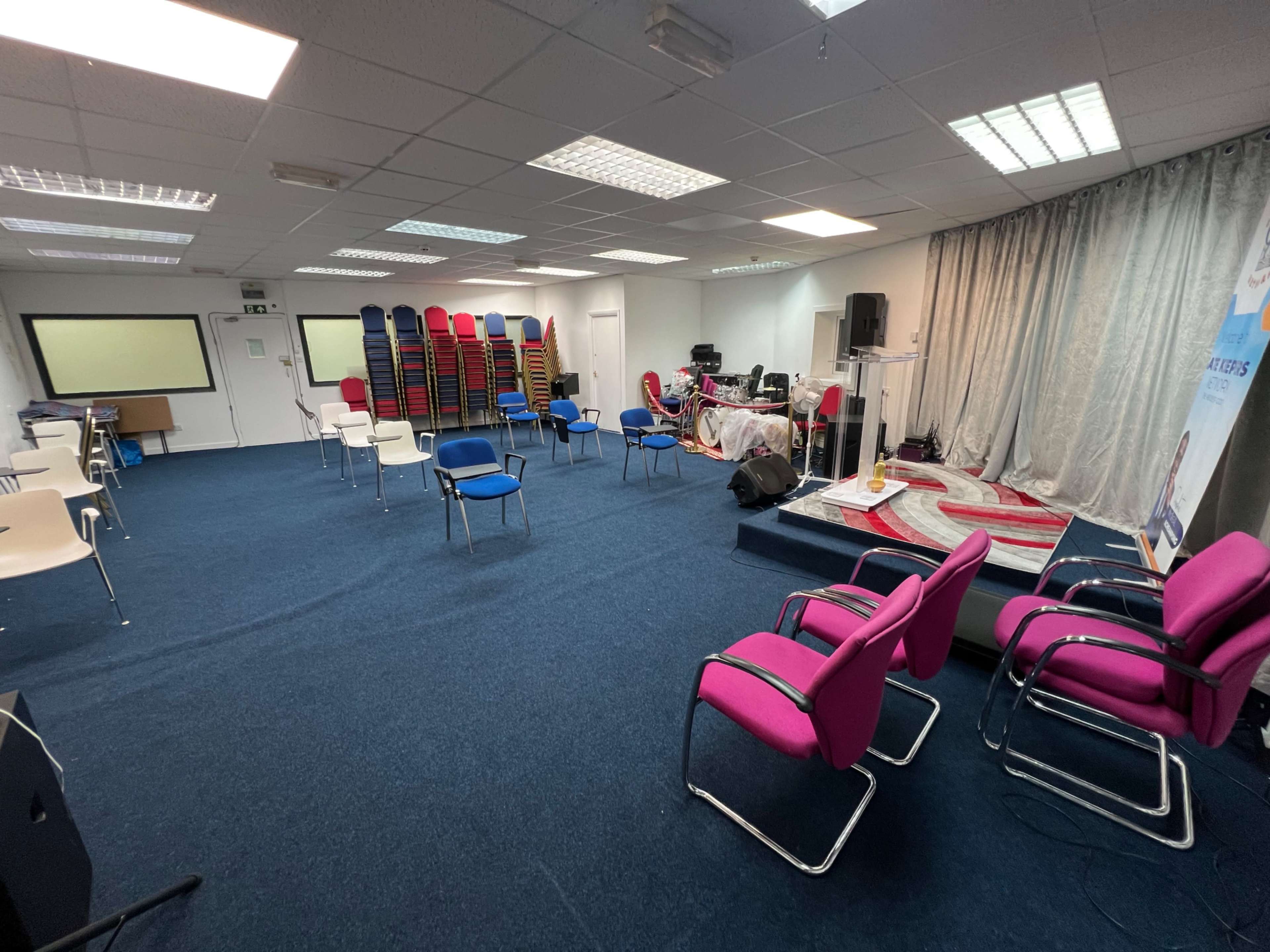 The image shows a conference room arranged with chairs in various colors and stacks of additional chairs against the wall, with a stage setup at the front.