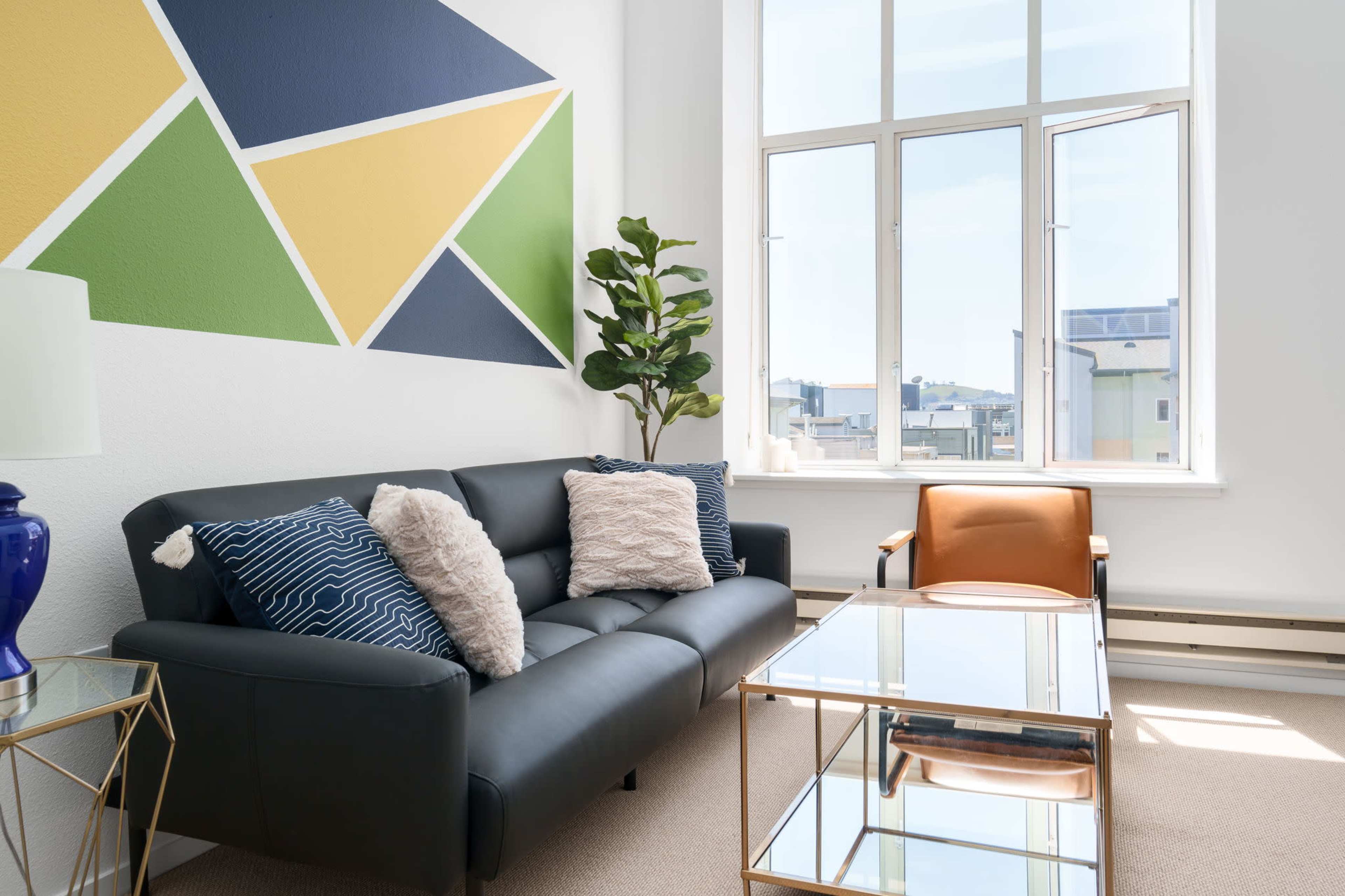 A modern living room featuring a black sofa with decorative pillows, a geometric wall mural, a potted plant, and a glass coffee table beside an orange accent chair.