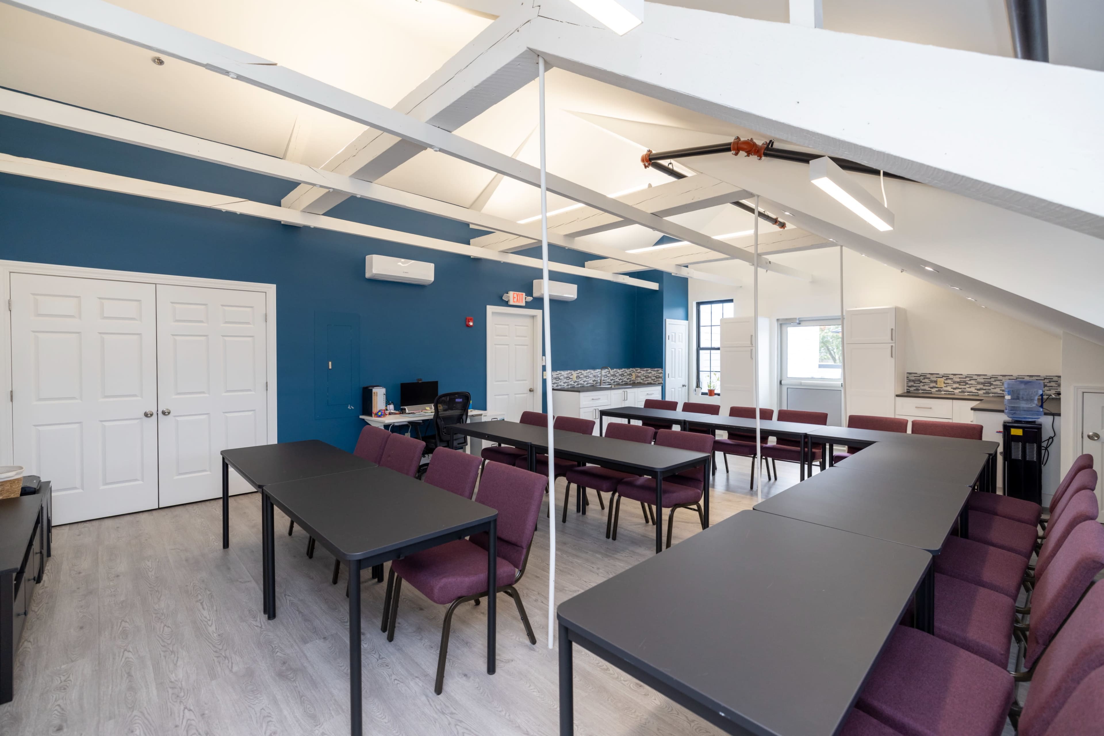 A bright, spacious conference room with several tables arranged in a U-shape and chairs positioned around them, featuring blue walls and large windows.