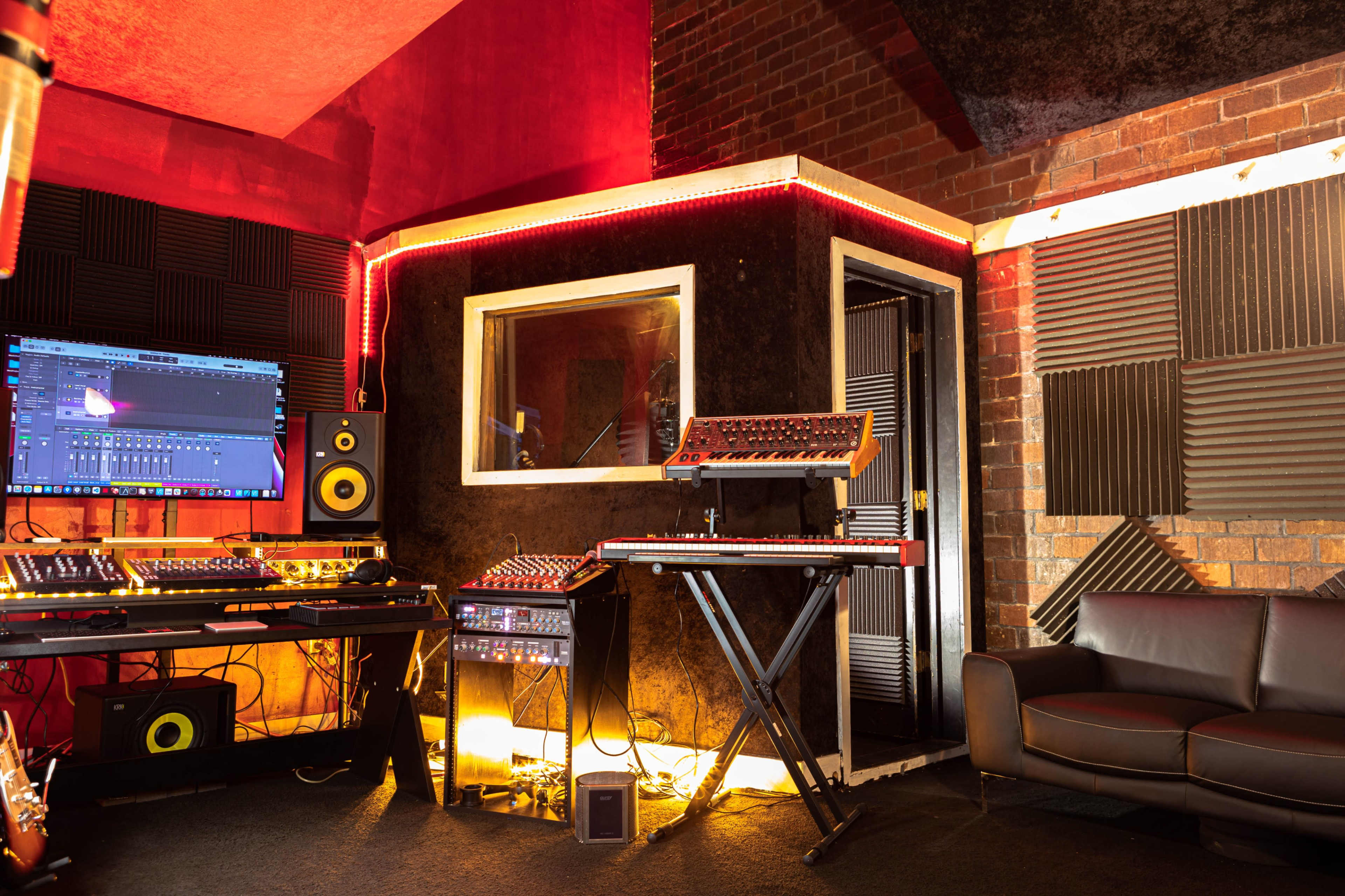 A music production studio equipped with various synthesizers, a computer monitor displaying audio software, and a comfortable seating area.