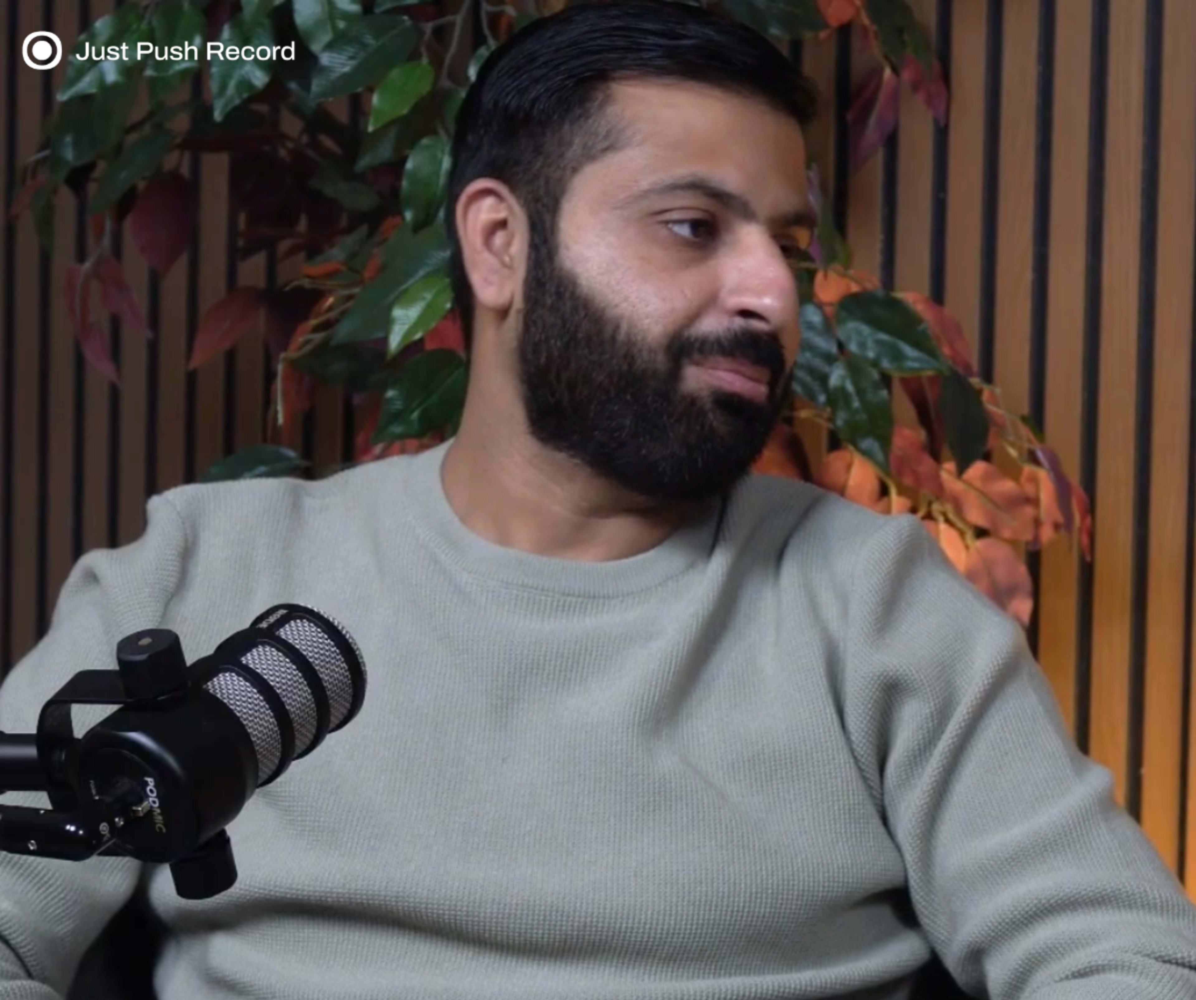 A man with a beard is seated and facing slightly to the side, while a microphone is positioned in front of him and decorative plants are visible in the background.