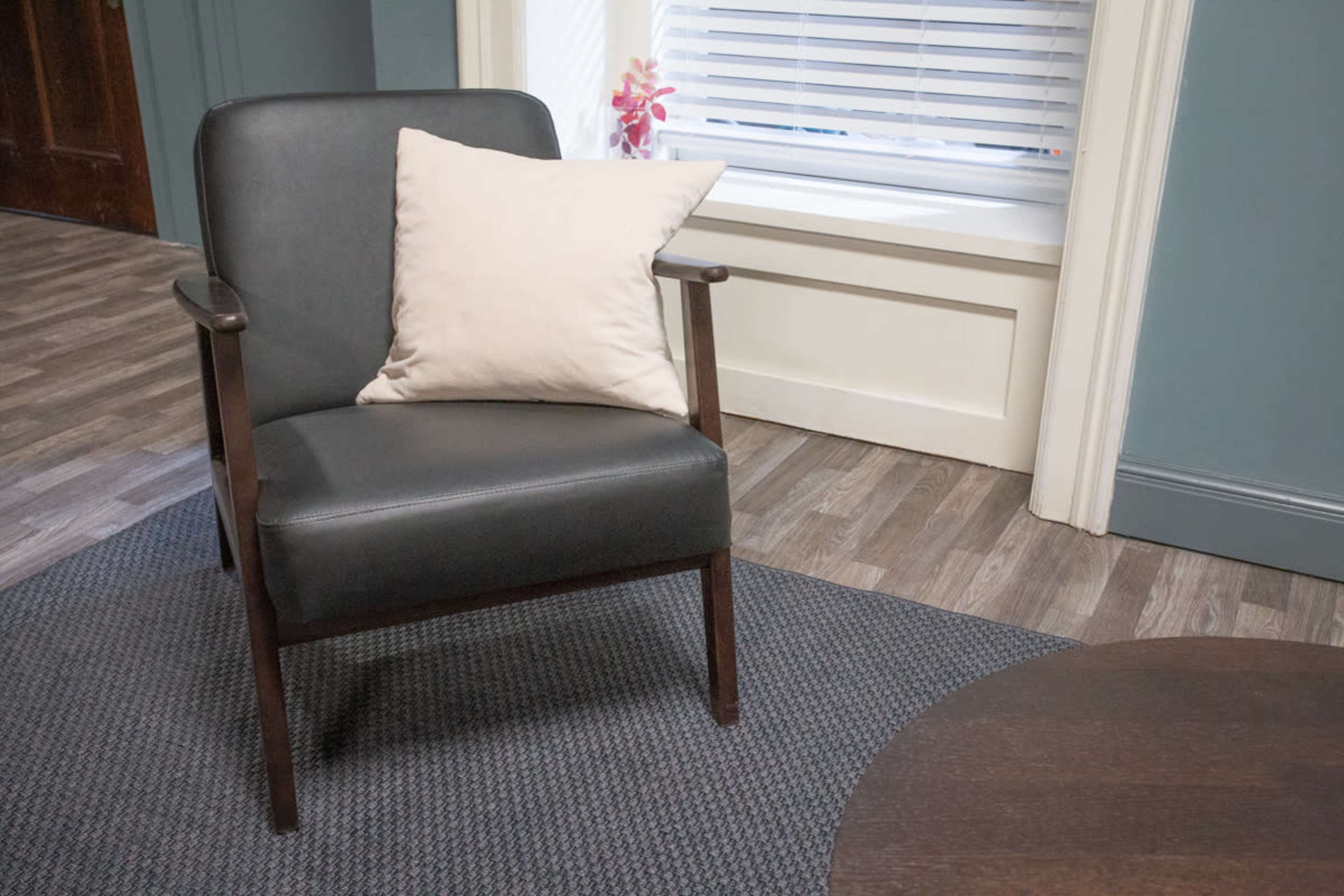 A black upholstered armchair with a light beige pillow sits beside a round wooden table in a room with blue walls and a window.