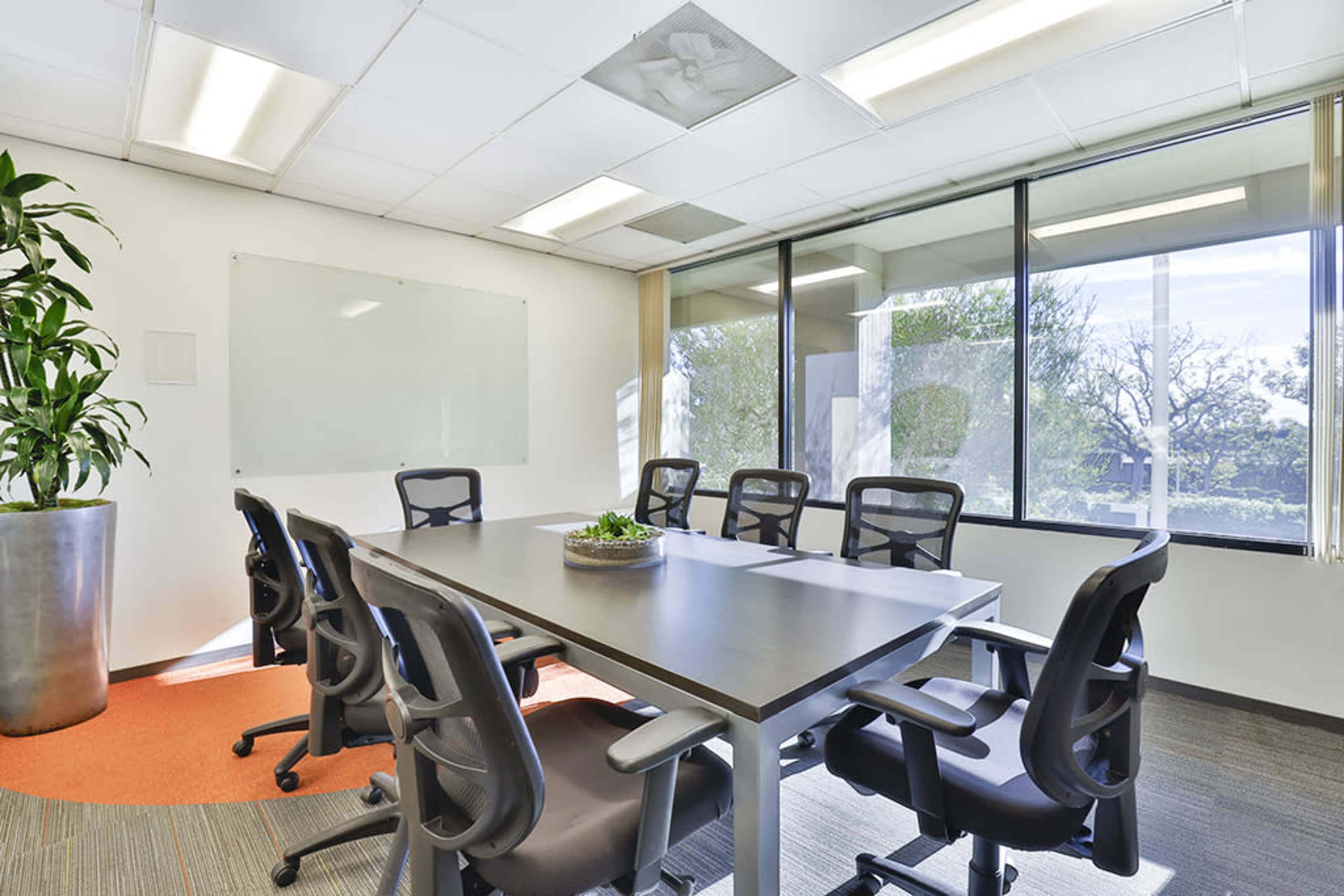 A modern conference room features a large table surrounded by ergonomic chairs, with a glass wall offering views of the outdoors and a plant in the corner.
