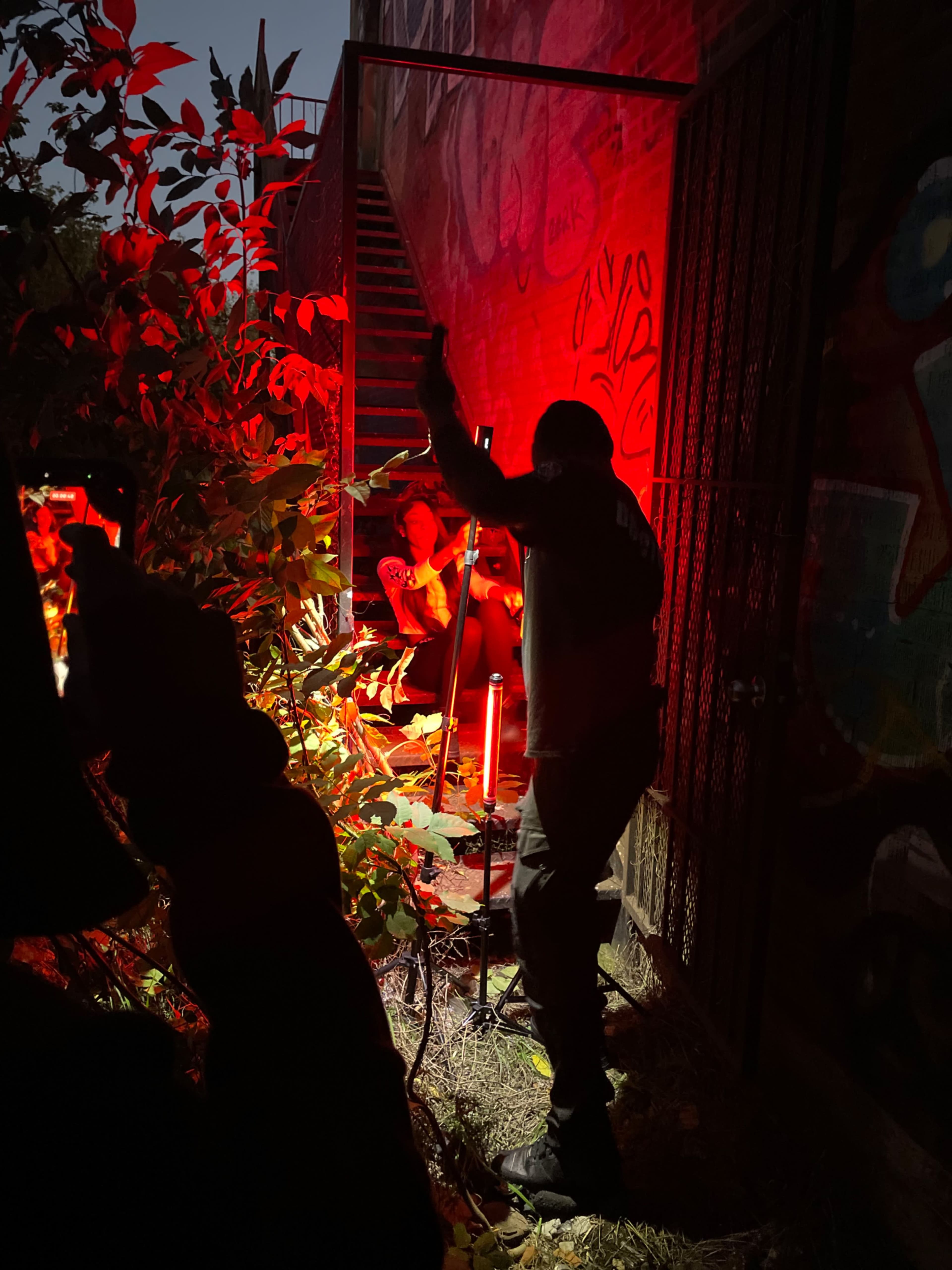 A person stands near bright red lights in an outdoor setting with graffiti-covered walls and a staircase in the background.