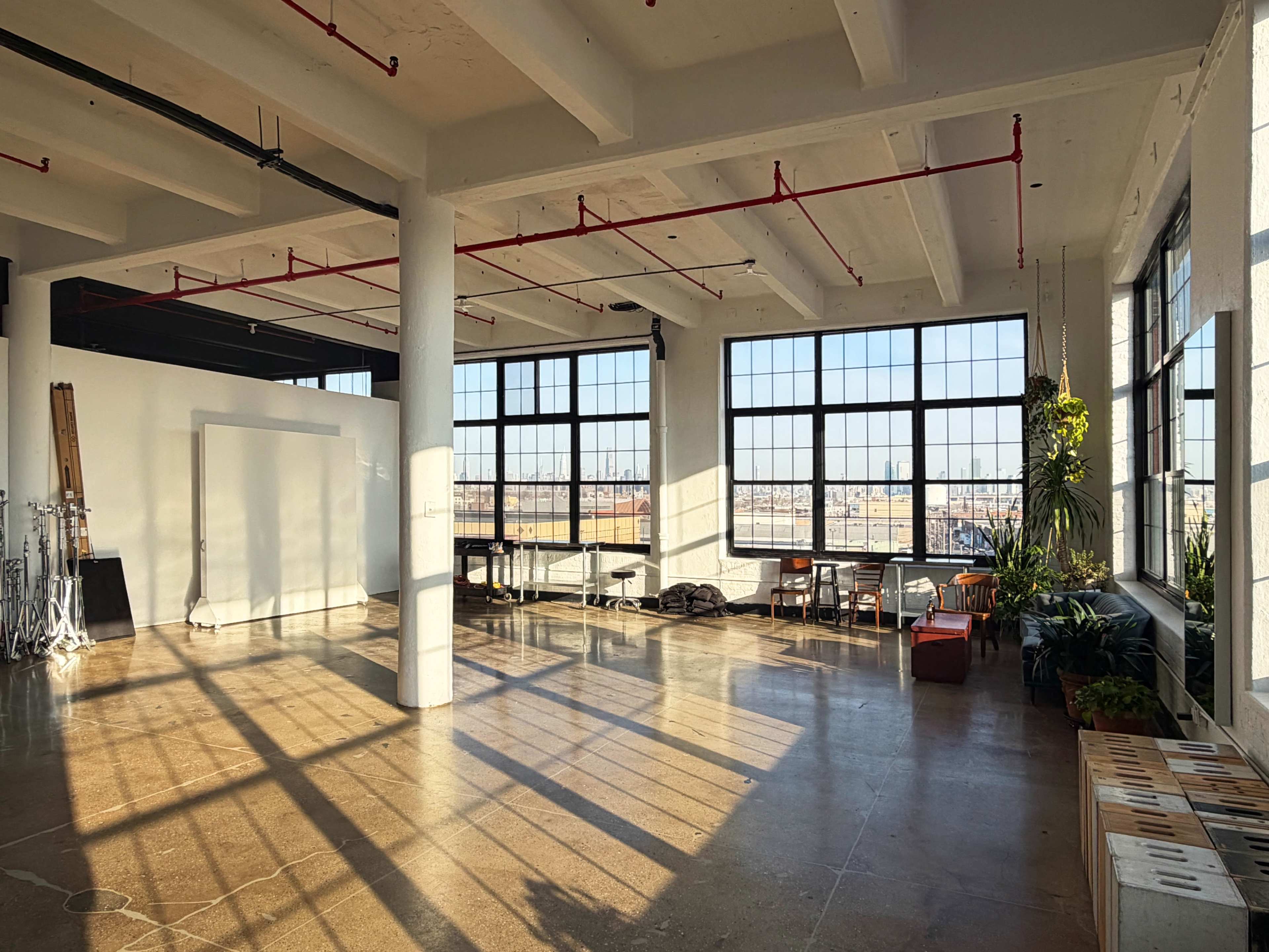 The image shows an industrial-style workspace with large windows, polished concrete floors, and plants in the corners, featuring a mix of furniture and lighting equipment.