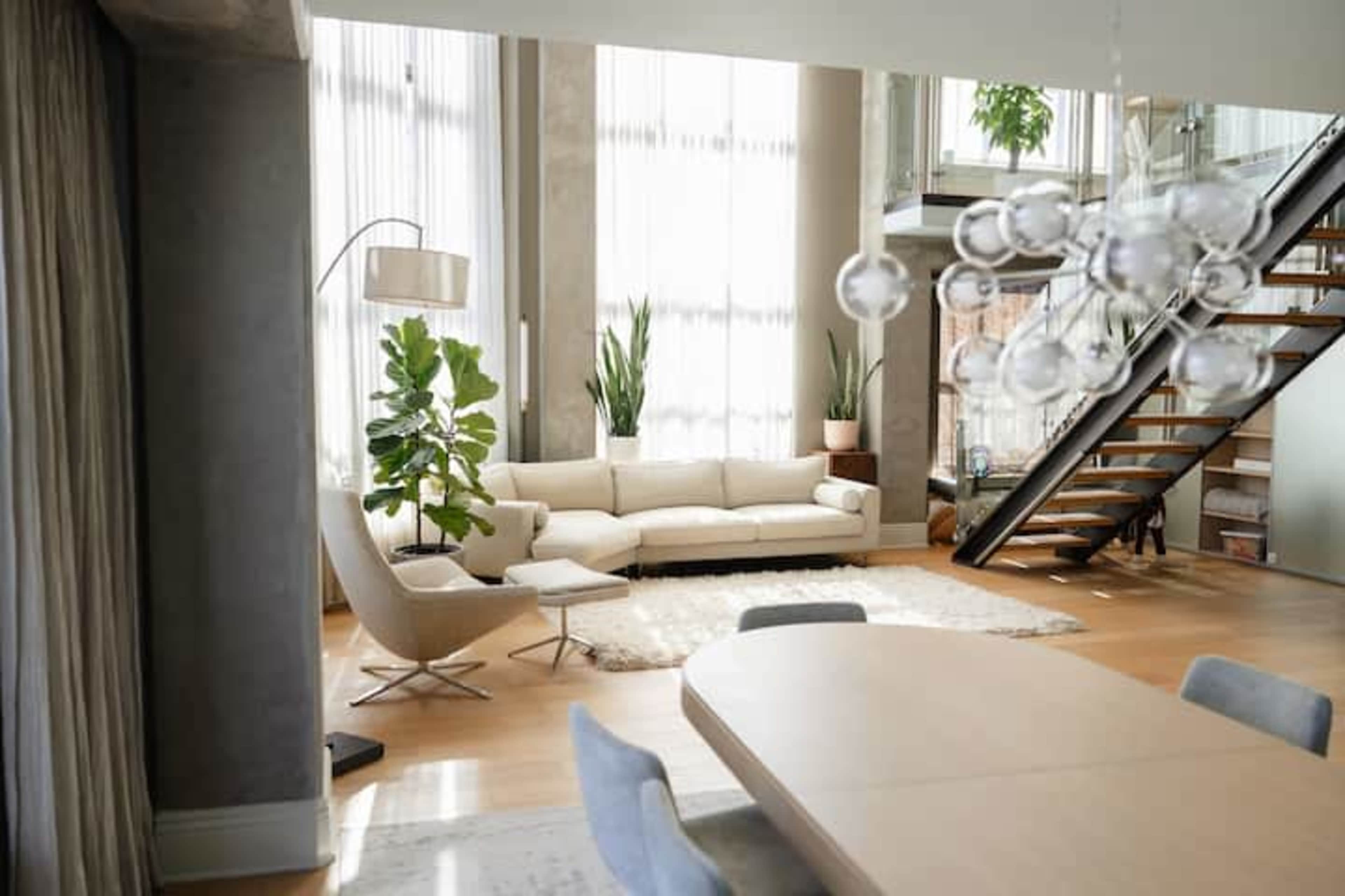The image shows a modern living space featuring a white sofa, a large rug, several indoor plants, and a dining area with a wooden table, accompanied by a staircase in the background.