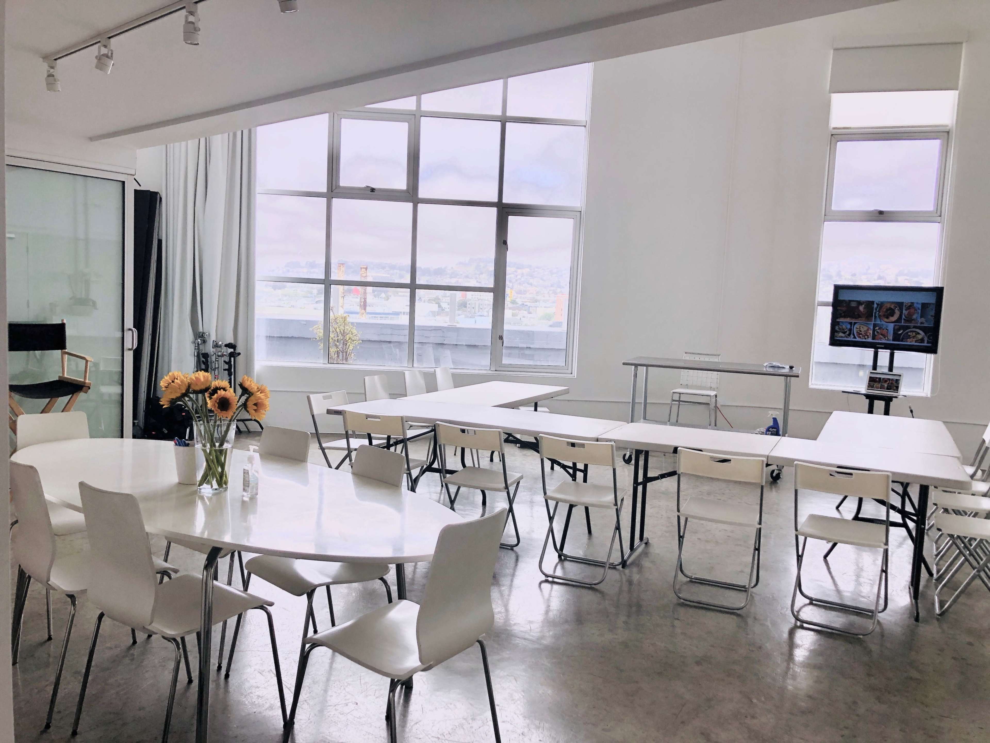 A spacious room features several white tables arranged with chairs and large windows offering a view of the city.