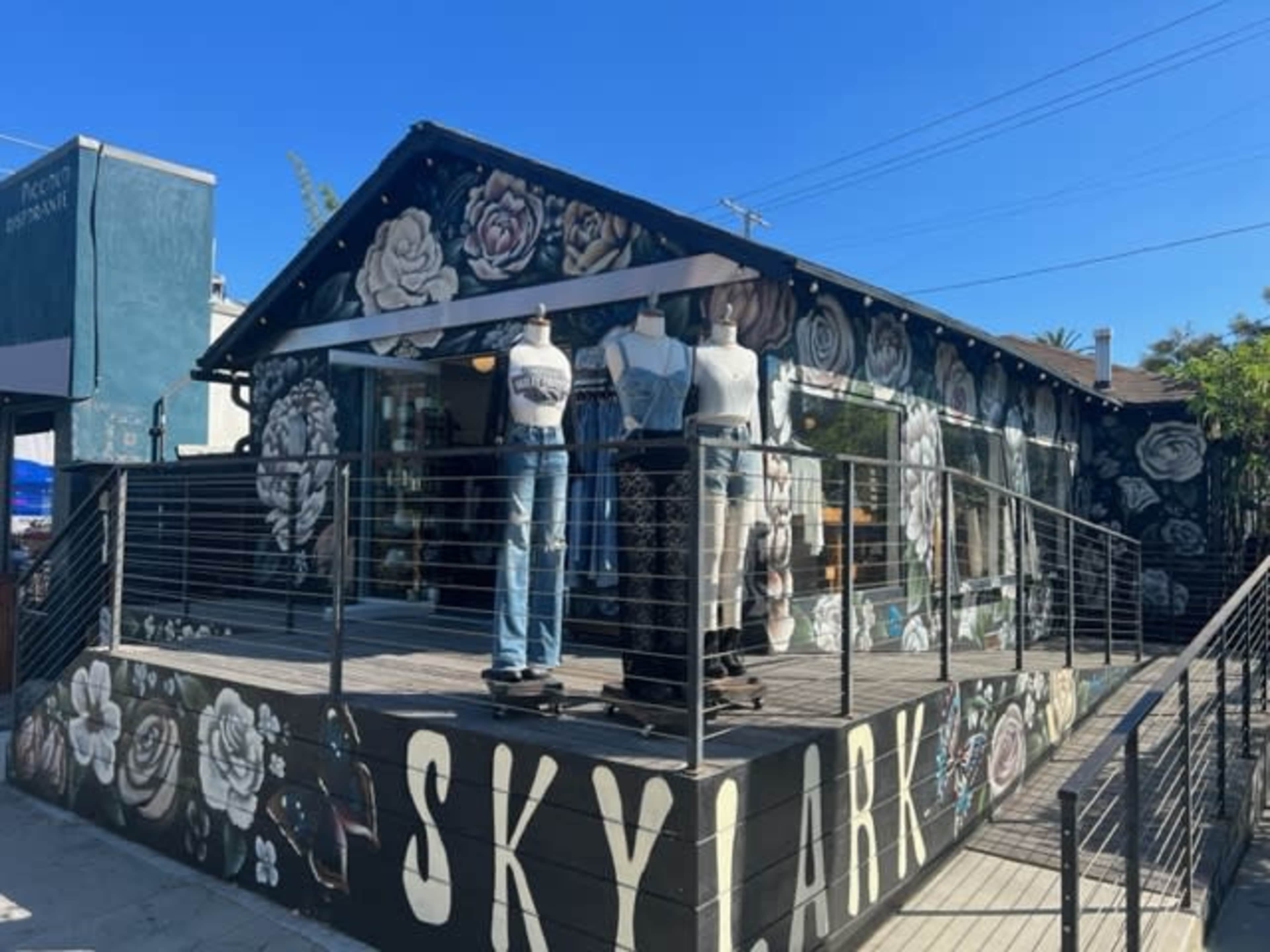 The image shows a retail shop called "SKYLARK" featuring a floral mural on its exterior and mannequins displaying clothing on a deck.