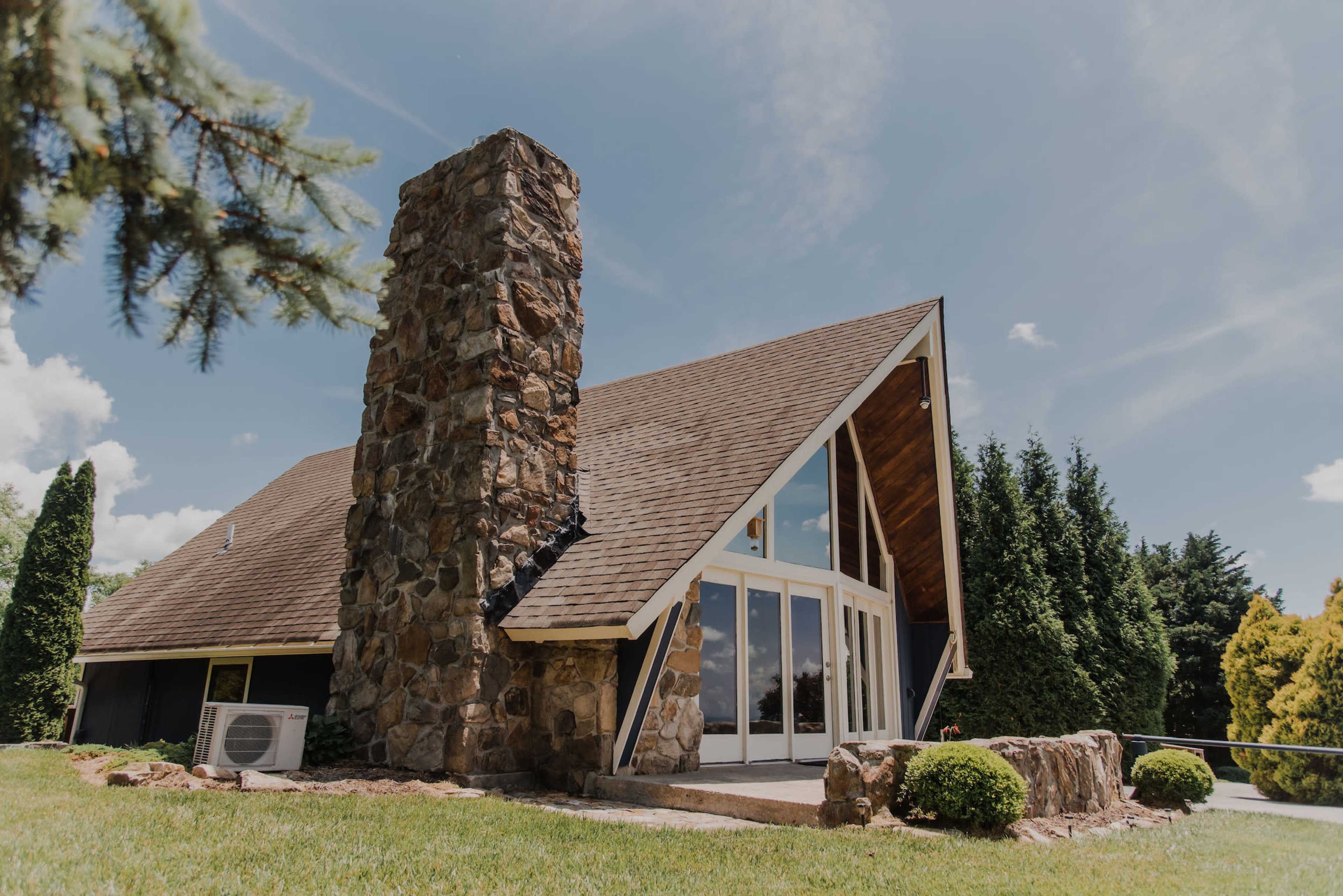 A modern house with a stone chimney, large windows, and a sloped roof, surrounded by green grass and trees.