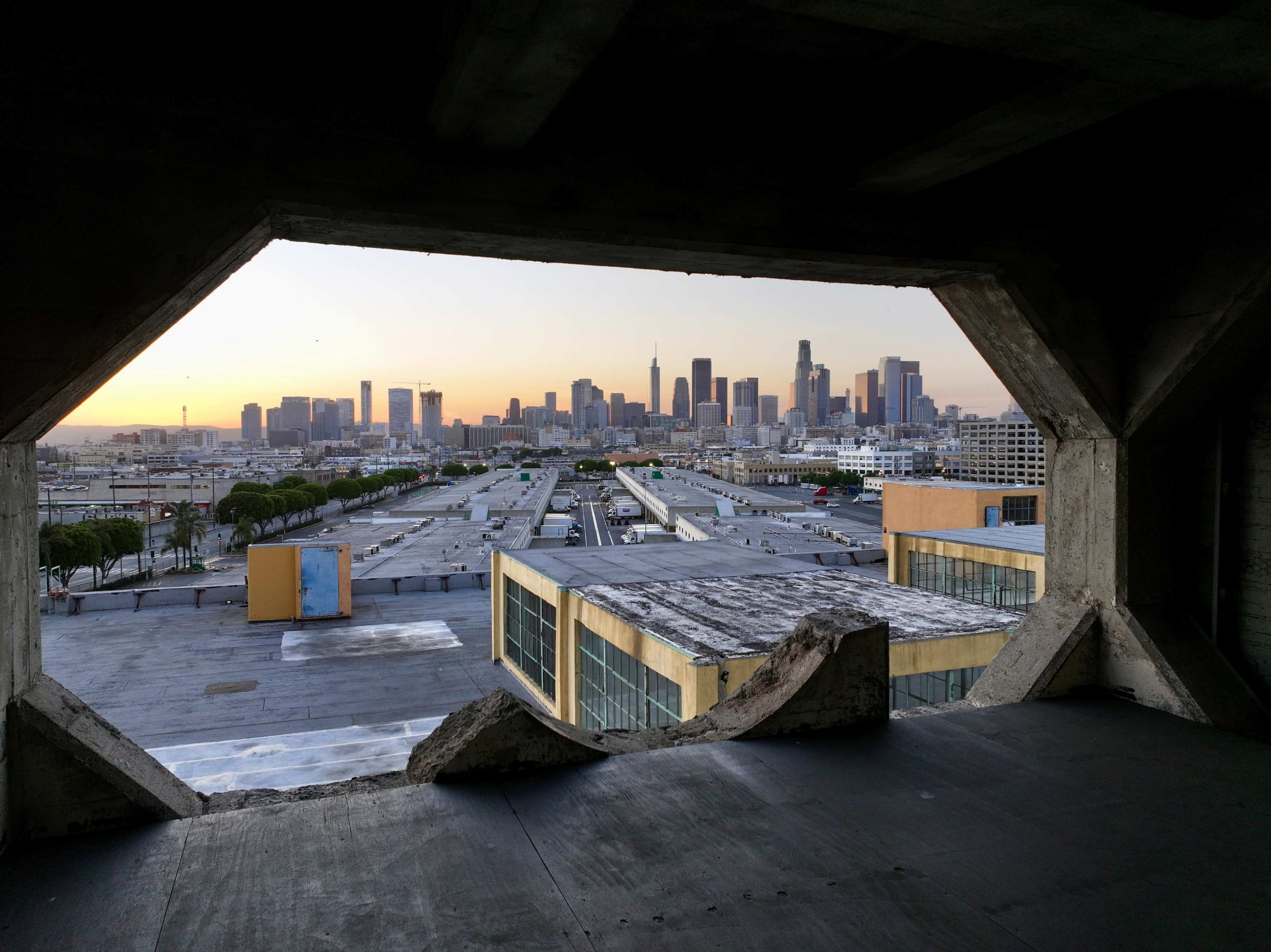 The photo shows a city skyline with tall buildings viewed through a broken concrete opening at sunset.