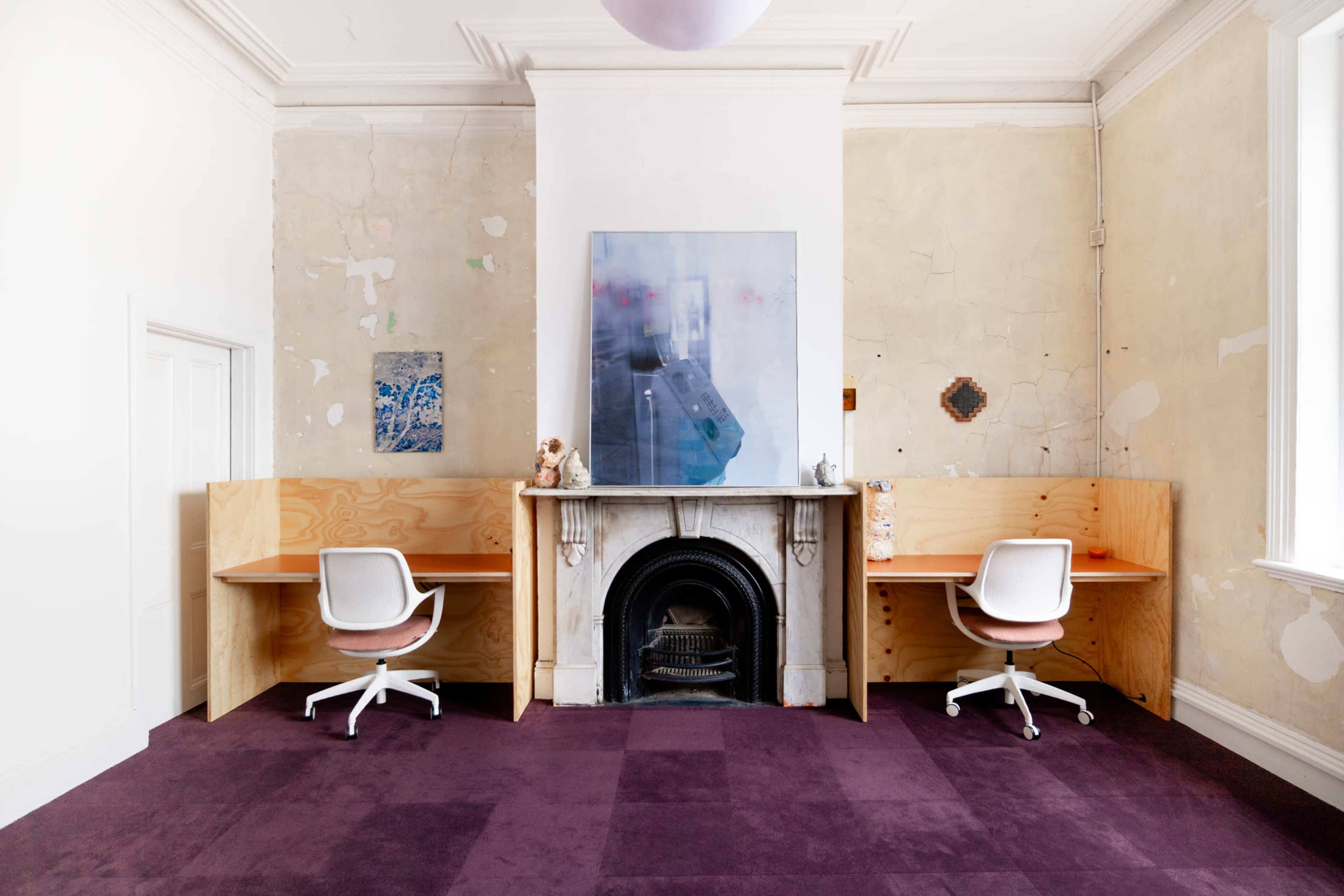 The image shows a room with two desks arranged on either side of a fireplace, featuring a large piece of artwork on the wall and purple carpet.