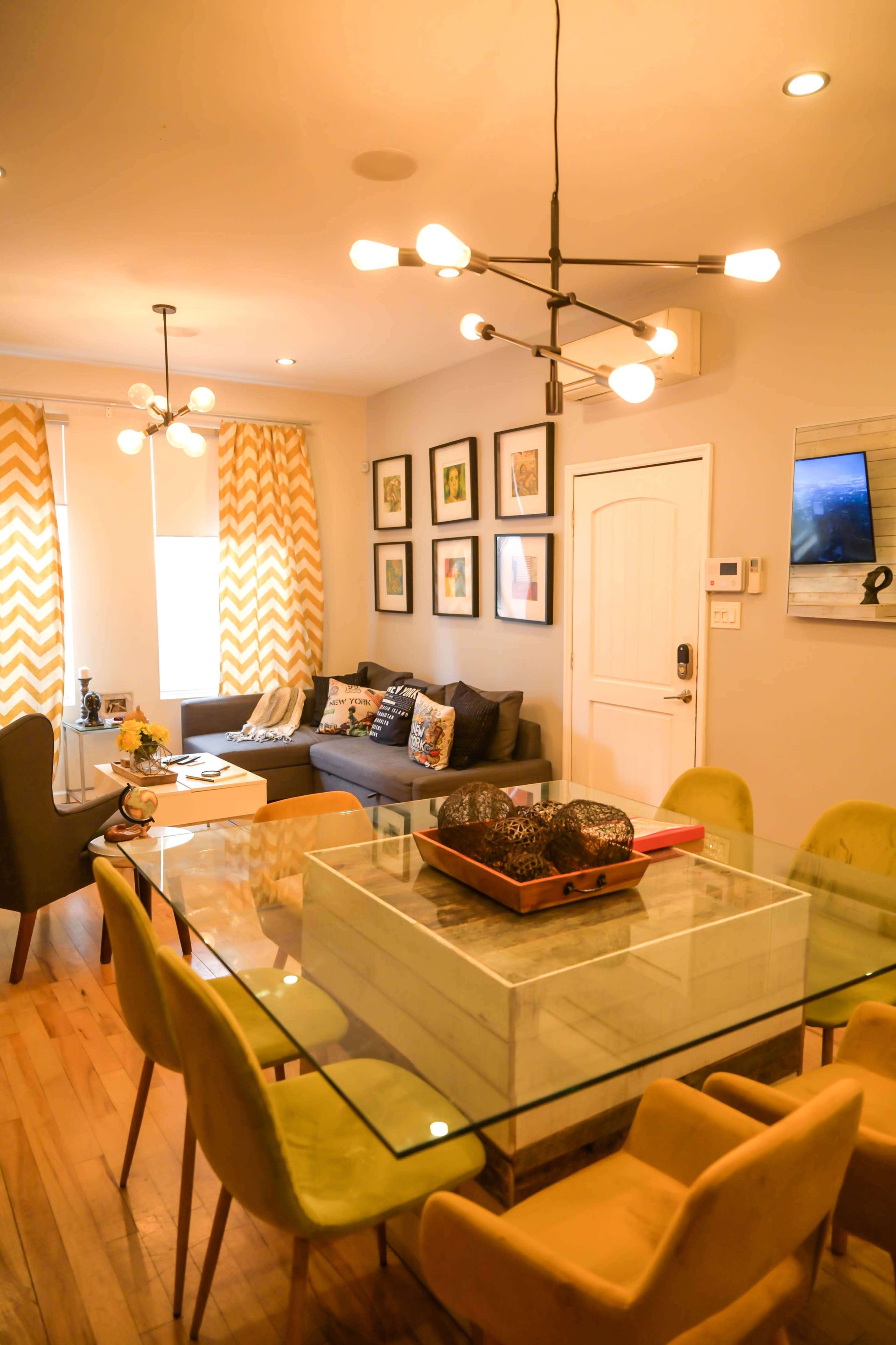 A modern living room and dining area with a glass dining table surrounded by upholstered chairs, a sectional sofa, framed artwork on the walls, and decorative lighting fixtures.